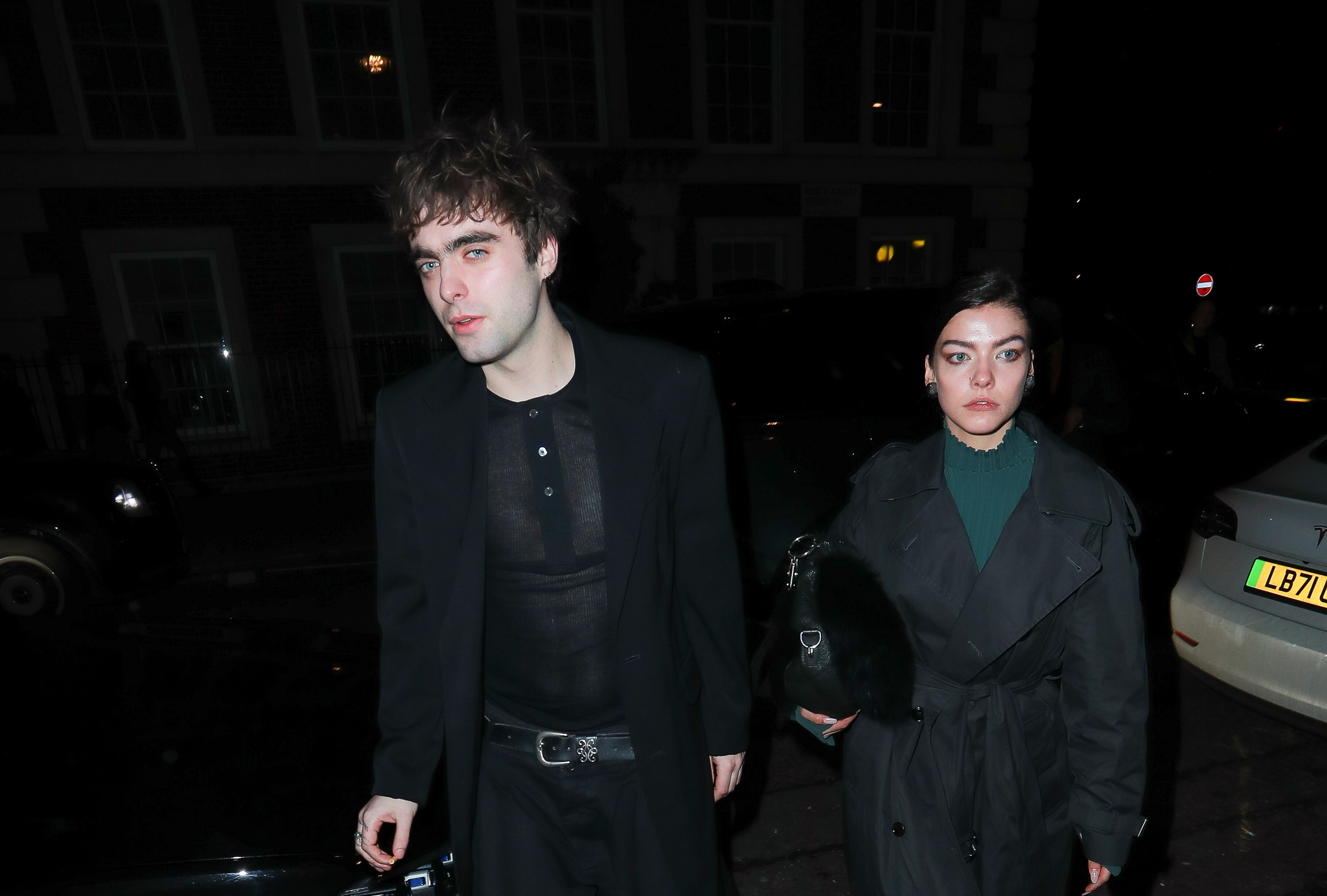 Lennon Gallagher and Isobel Richmond walking together at night, attending the Burberry Winter 2024 show after party during London Fashion Week. Lennon wears an all-black ensemble with a sheer top under a blazer, while Isobel sports a green turtleneck under a black trench coat.