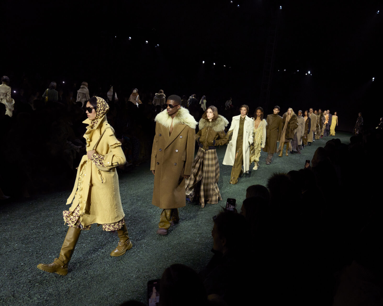 Models walk the runway in a variety of layered winter ensembles from Burberry's Winter 2024 collection, with an emphasis on oversized coats and rich textures, against a backdrop of a dimly lit, green-floored runway surrounded by an audience.