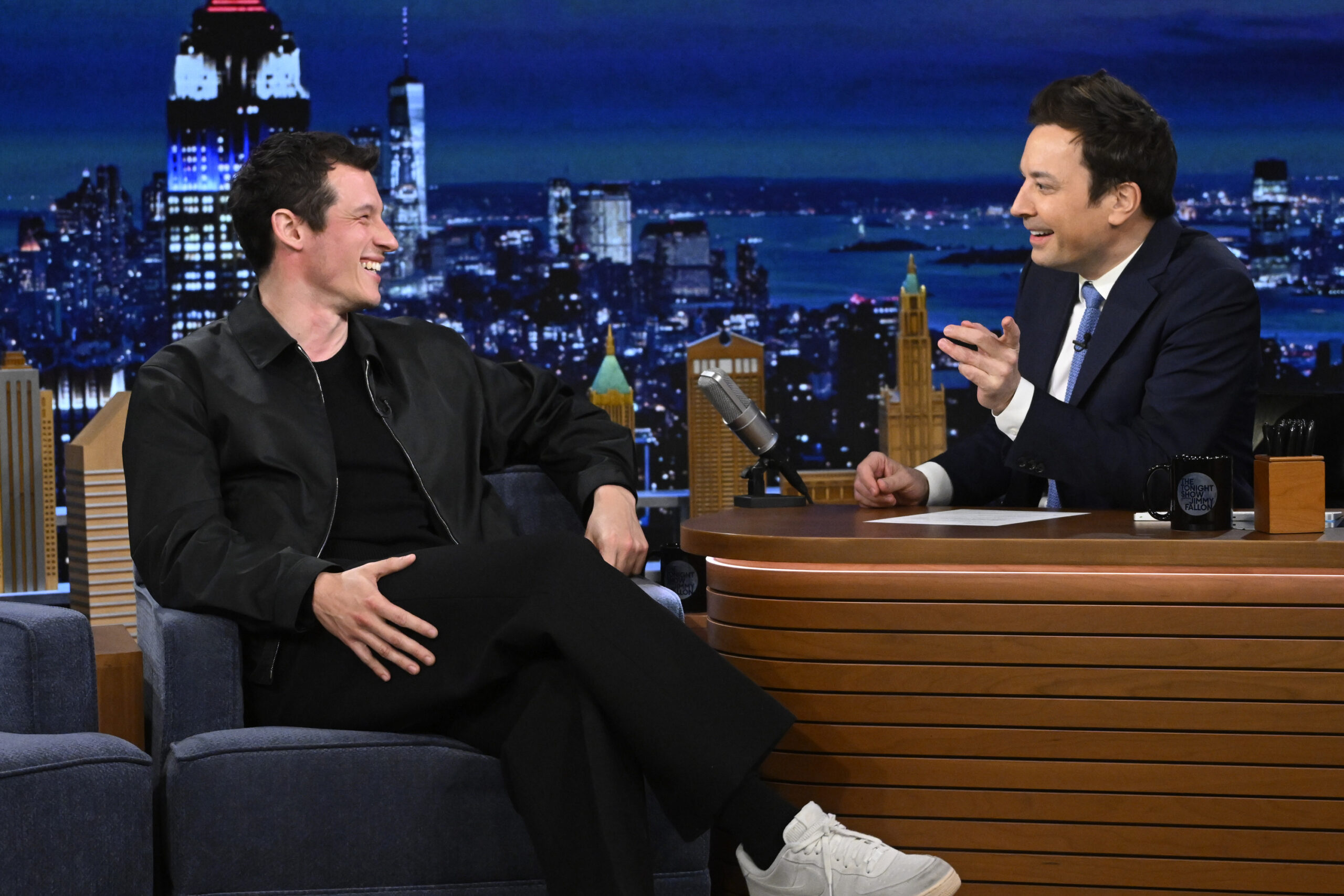 Actor Callum Turner and Jimmy Fallon engaged in a lively conversation on a talk show set, with one man seated across from the host, both laughing and gesturing.