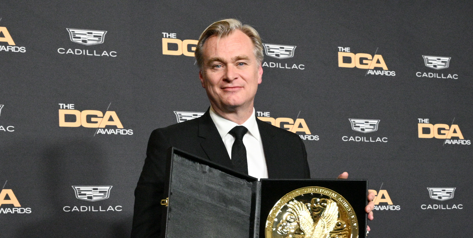 Christopher Nolan, dressed in a formal black suit, holding an award at the 76th Annual DGA Awards.