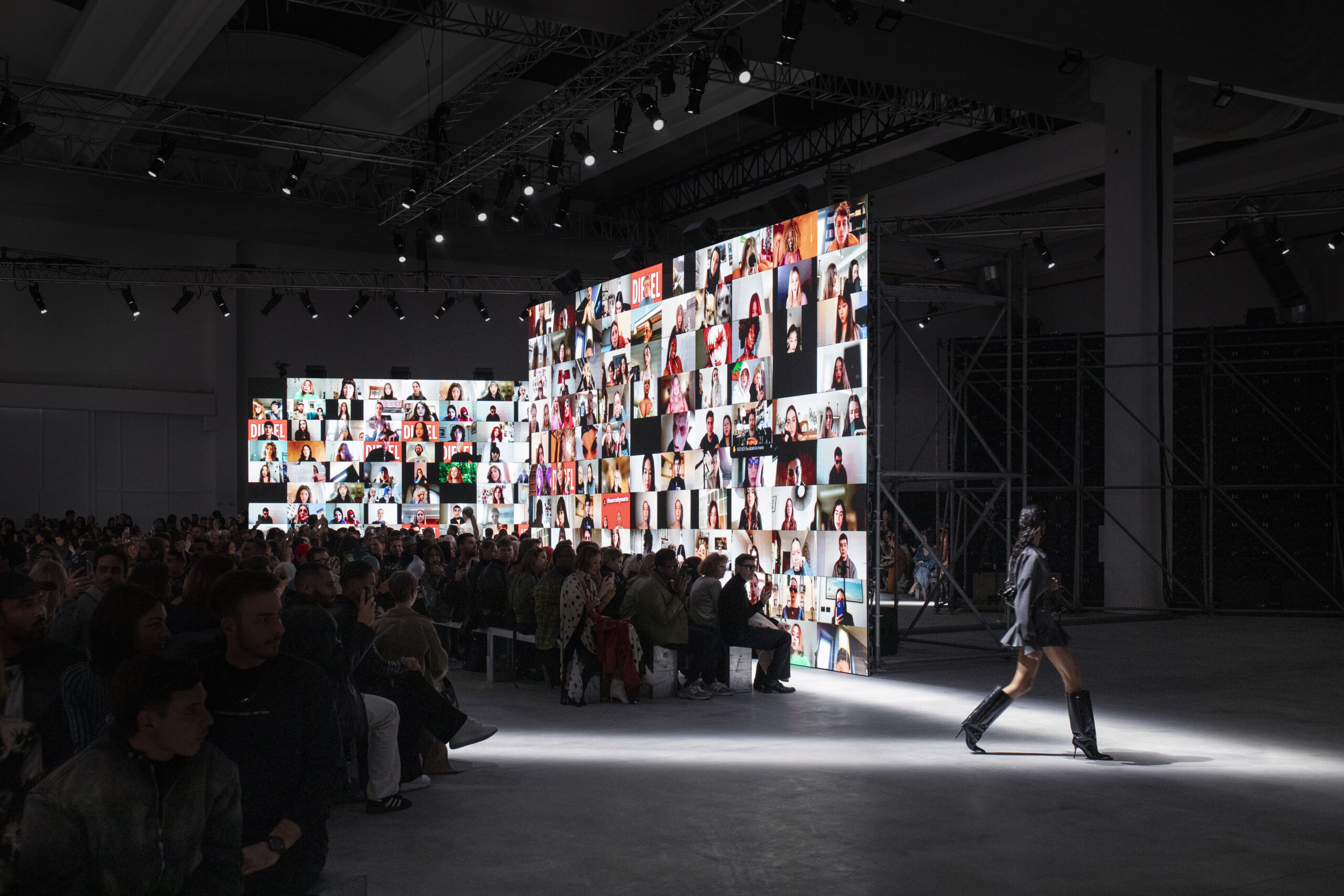 A fashion show setting featuring a large backdrop with numerous live video calls showcasing Diesel fans from around the world, as a model walks the runway at Diesel's Fall Winter 2024 collection during Milan Fashion Week.