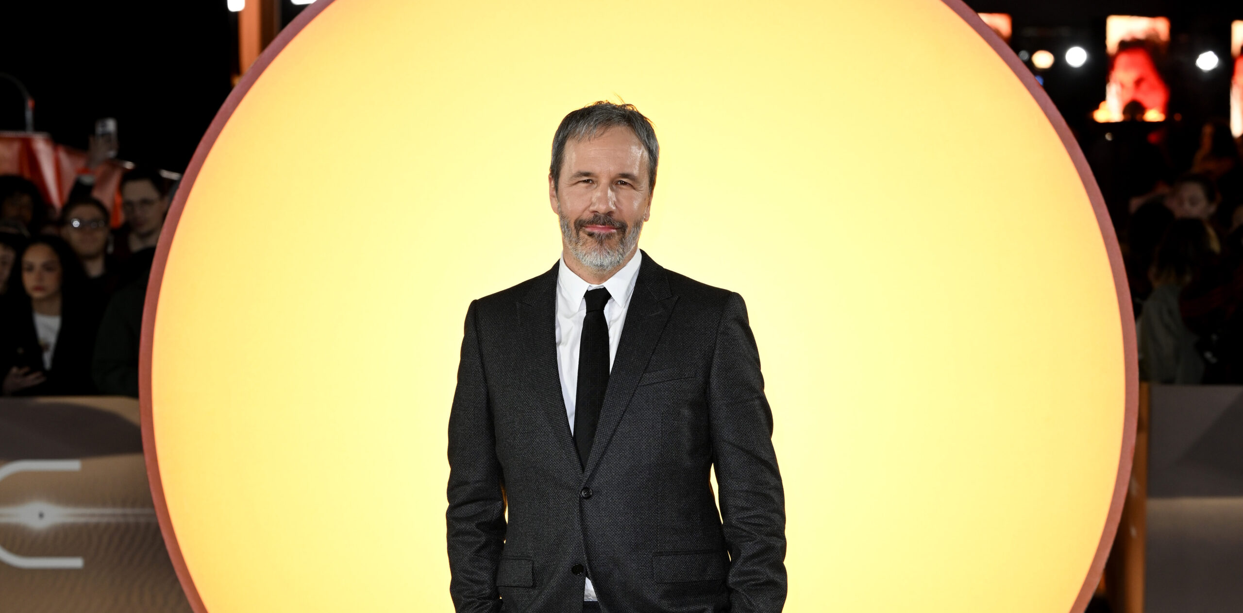 Denis Villeneuve standing confidently before a large, glowing orb at the London premiere of 'Dune: Part Two,' wearing a Dior gray wool peak lapel suit, with a black silk tie, white cotton shirt, and black leather derbies.