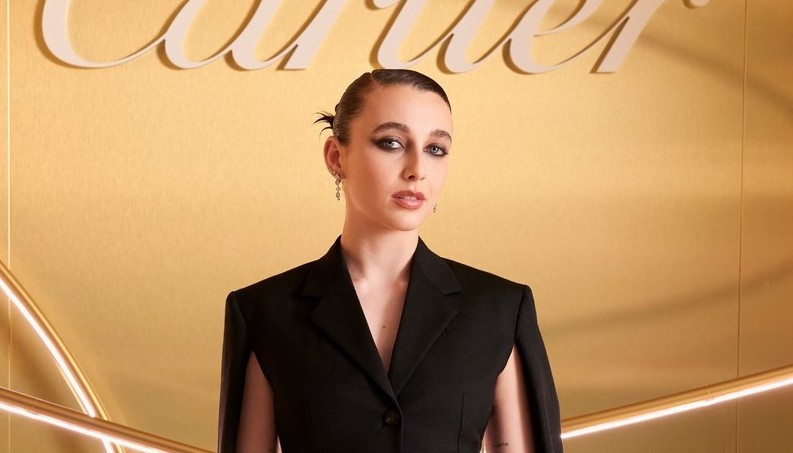 Emma Chamberlain poses at the Cartier event, dressed in a Ferragamo black tailored jacket with unique slashes at the under-arms, and matching flare trousers. She accessorizes with a small black handbag and delicate jewelry, standing before a golden background with the Cartier logo illuminated above.