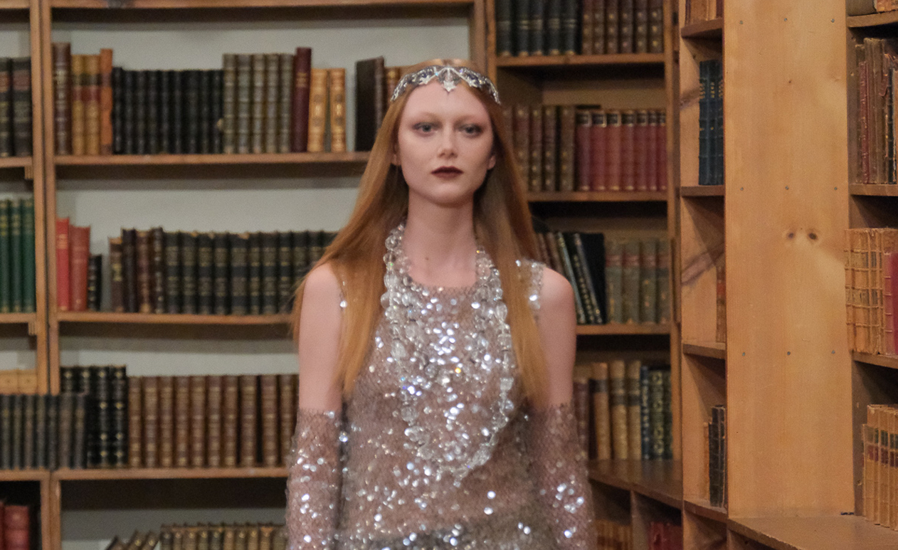 A model strides between bookshelves in a glittering Anna Sui Fall/Winter 2024 garment, with a shimmering sleeveless top and sequined pants, crowned by a bejeweled headpiece.