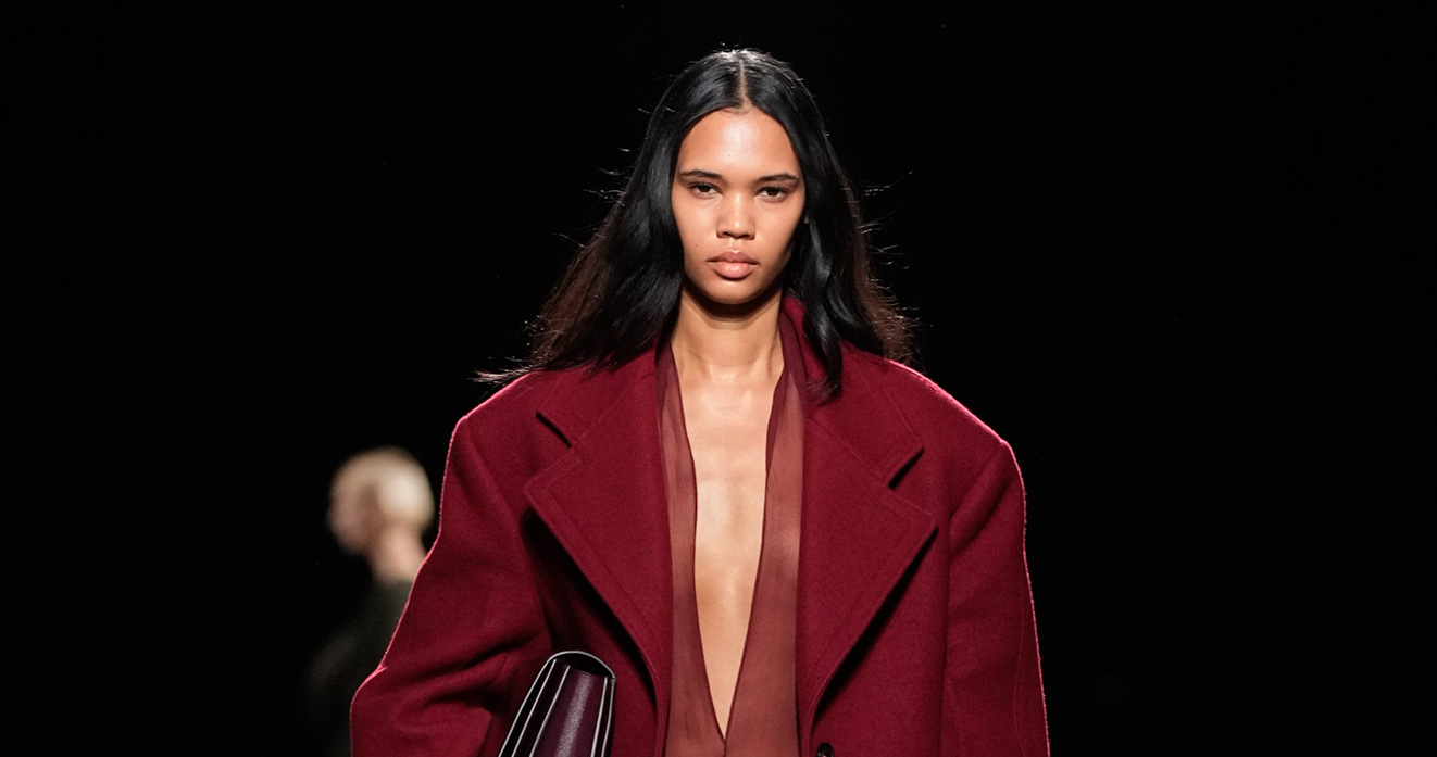 A model walks the runway in an oversized burgundy coat with a plunging neckline, embodying the Ferragamo Fall Winter 2024 collection's theme of bold self-expression and elegance. The coat's rich color and tailored structure showcase the collection's nod to the luxurious and liberated style of the Roaring Twenties, updated with a modern twist for contemporary appeal.