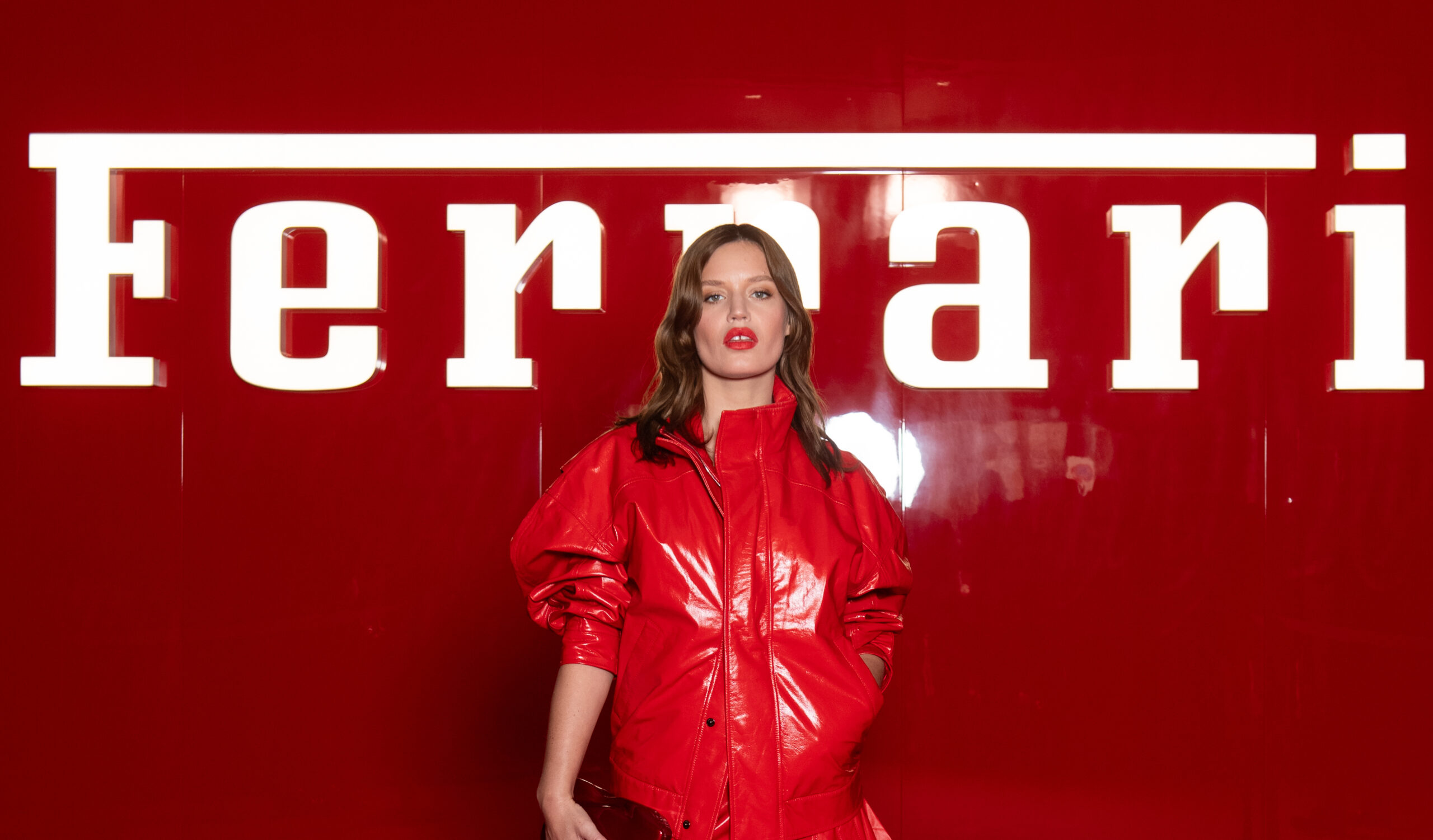 Georgia May Jagger stands confidently in front of a bold, red Ferrari backdrop, the brand name prominent above her head. She wears a vibrant red, shiny leather jacket that radiates luxury and style, complementing the backdrop and hinting at the fusion of high fashion and automotive excellence.