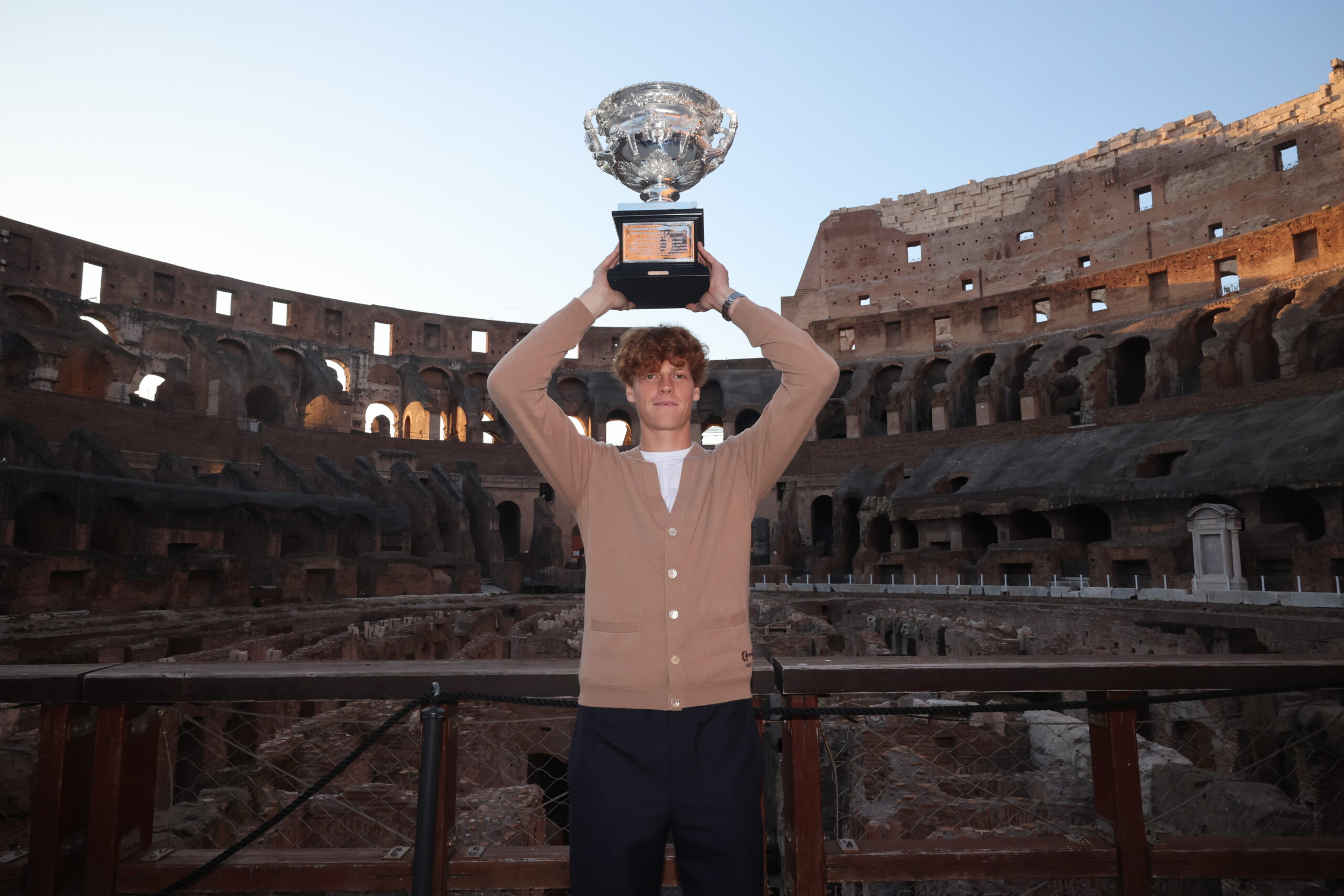 Tennis champion Jannik Sinner holds the Norman Brookes Challenge Cup aloft at the historic Colosseum in Rome, wearing a Gucci cashmere cardigan, celebrating his victory with classical grandeur in the background.