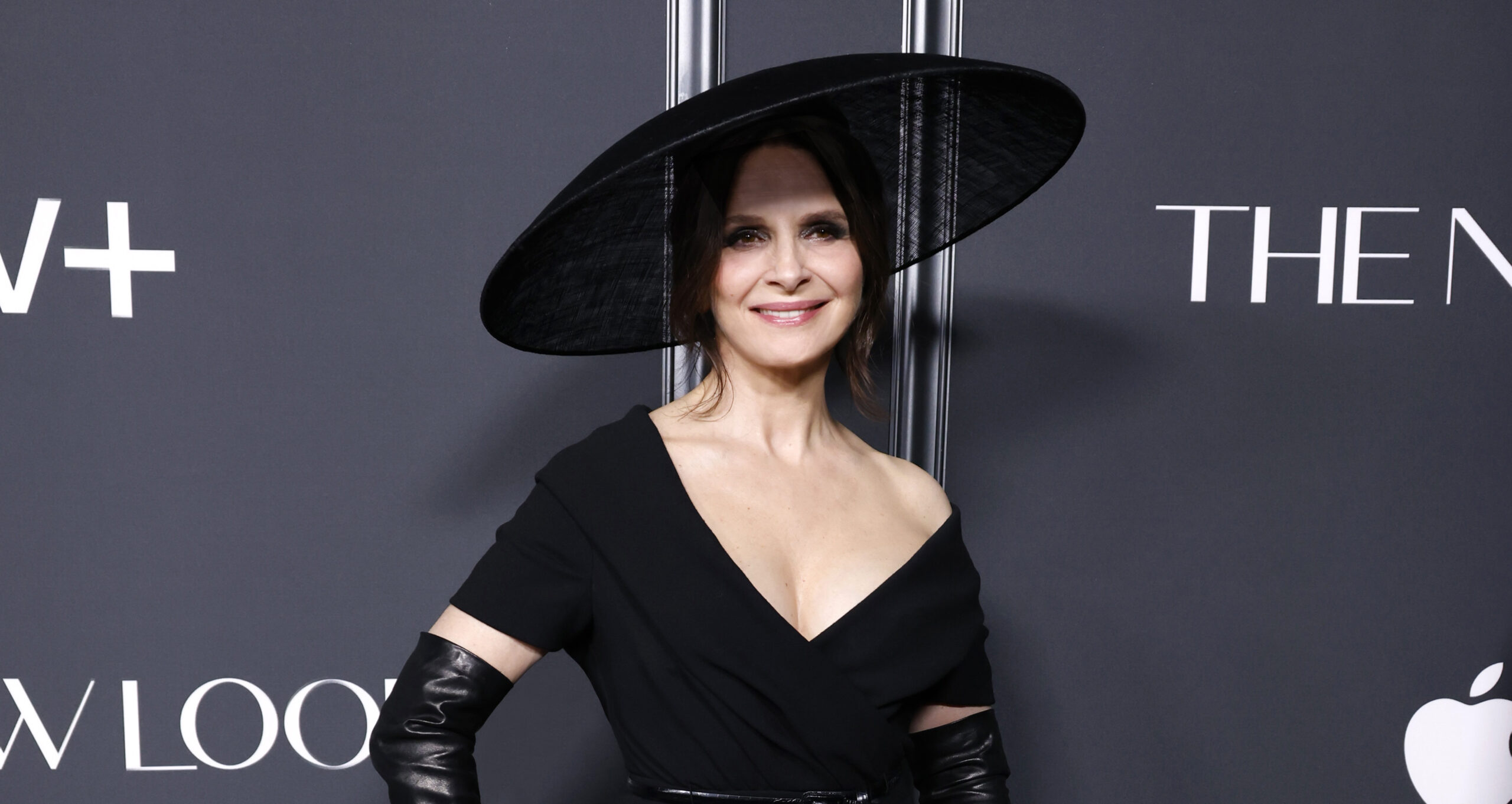 Juliette Binoche stands elegantly at the Apple TV+'s "The New Look" world premiere in New York City, wearing a classic Dior black wool crepe dress with a black sisal hat, leather gloves, and boots, symbolizing the timeless elegance of Dior.