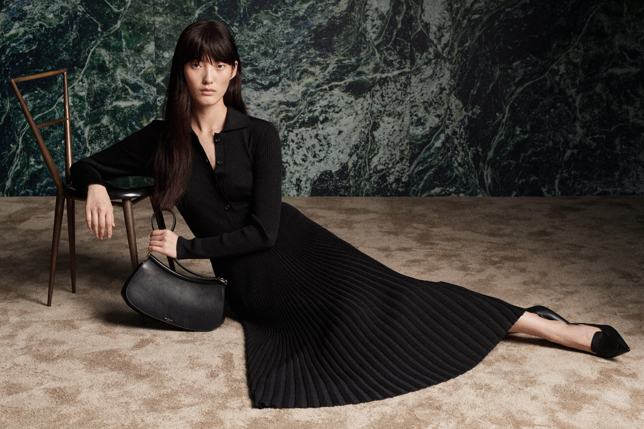 A woman lounges elegantly in a pleated black dress from Kiton's Fall/Winter 2024 Collection against a marbled green background, with a classic leather handbag by her side.