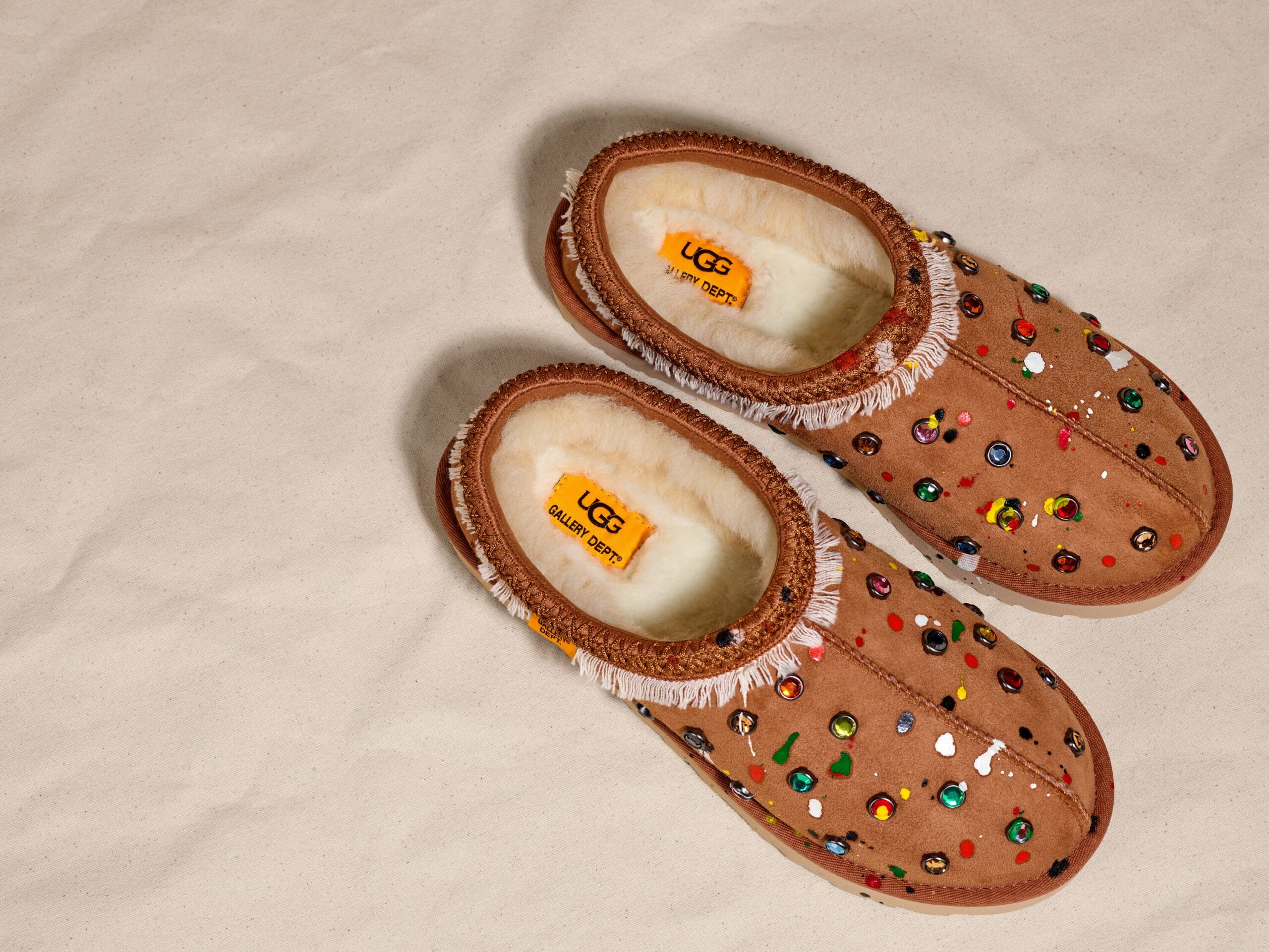 A pair of UGG x Gallery Dept. Tasman Slippers with chestnut suede adorned with colorful jewels and studs against a beige background.