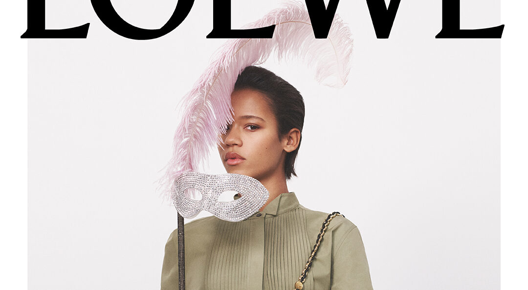 Taylor Russell in LOEWE's SS24 campaign, elegantly holding a crystal-embellished mask with pink feathers, wearing a green blouse, brown textured pants, and carrying a sleek black LOEWE bag
