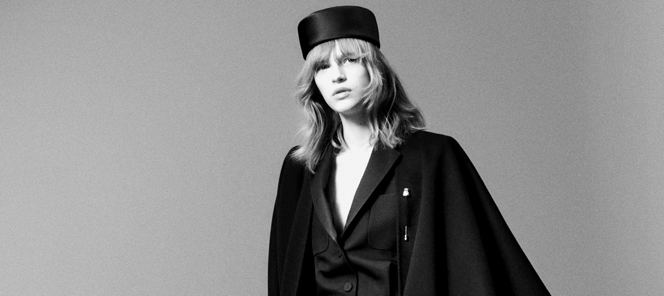 A monochromatic image of a model wearing Loro Piana's Fall/Winter 2024-2025 collection, showcasing a black blazer with a relaxed fit and a flat-topped, cylindrical hat. The model's expression is contemplative, and her hair falls in a casual style framing her face.
