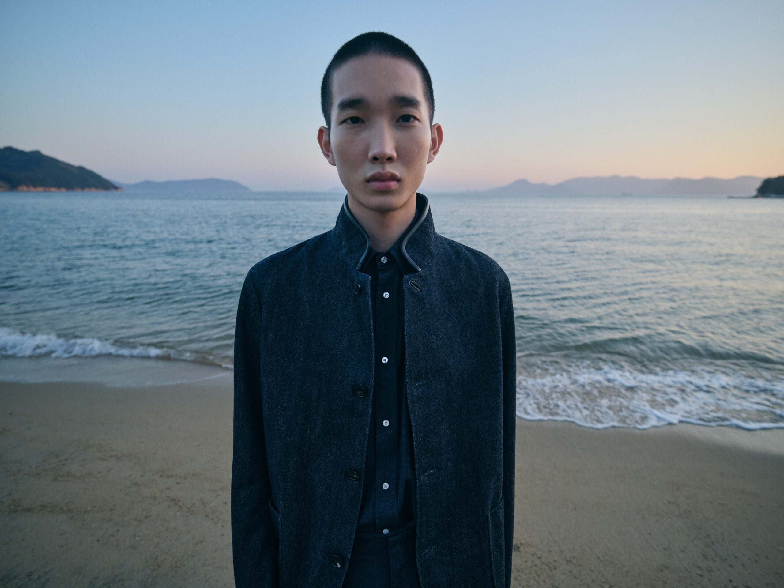 A man stands on a sandy beach at dusk, with gentle waves lapping at the shore and distant mountains under a soft sky. He is wearing the Loro Piana Spagna Jacket, a symbol of relaxed luxury with a distinctive standing collar, spacious patch pockets, and elegant horn buttons.