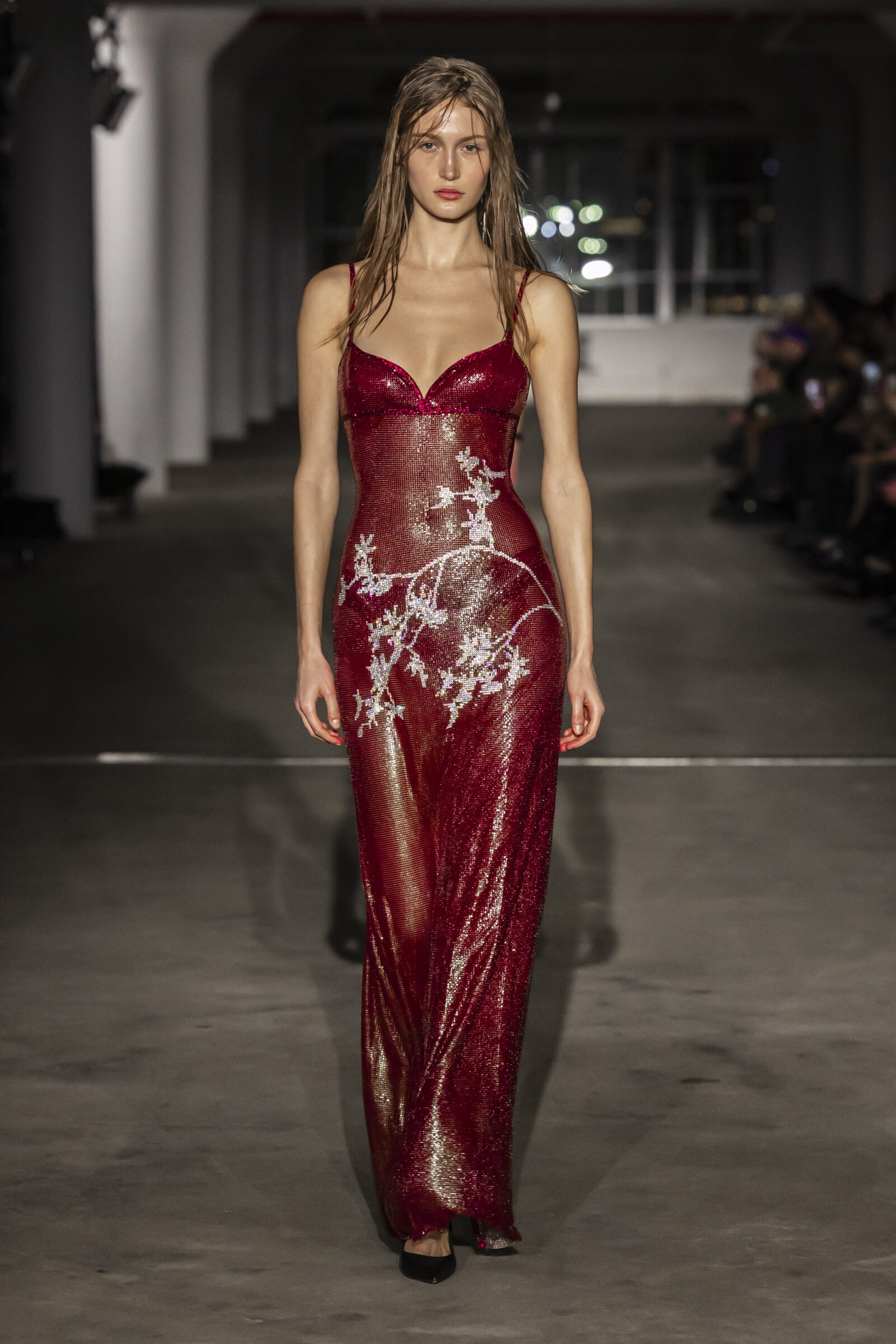 A model gracefully showcases a sparkling red sequined gown with delicate white floral accents, featuring a deep neckline and fitted silhouette, at Ludovic de Saint Sernin's AW24 collection show during New York Fashion Week.