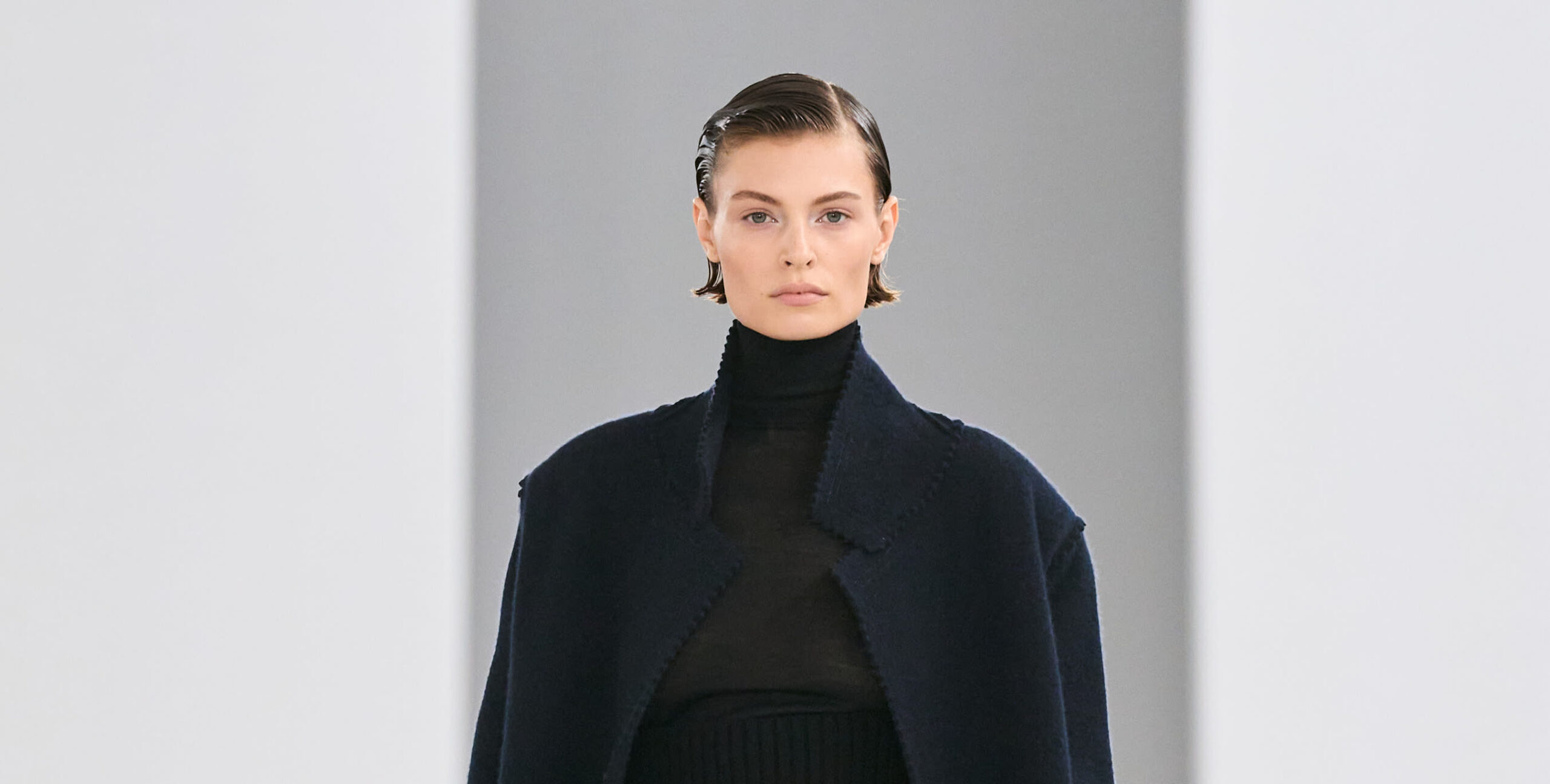 A portrait of a model at a fashion show wearing a dark blue cropped jacket and a high-neck sweater from Max Mara's Fall-Winter 2024 collection, styled with a sleek, combed-back hairdo and natural makeup.