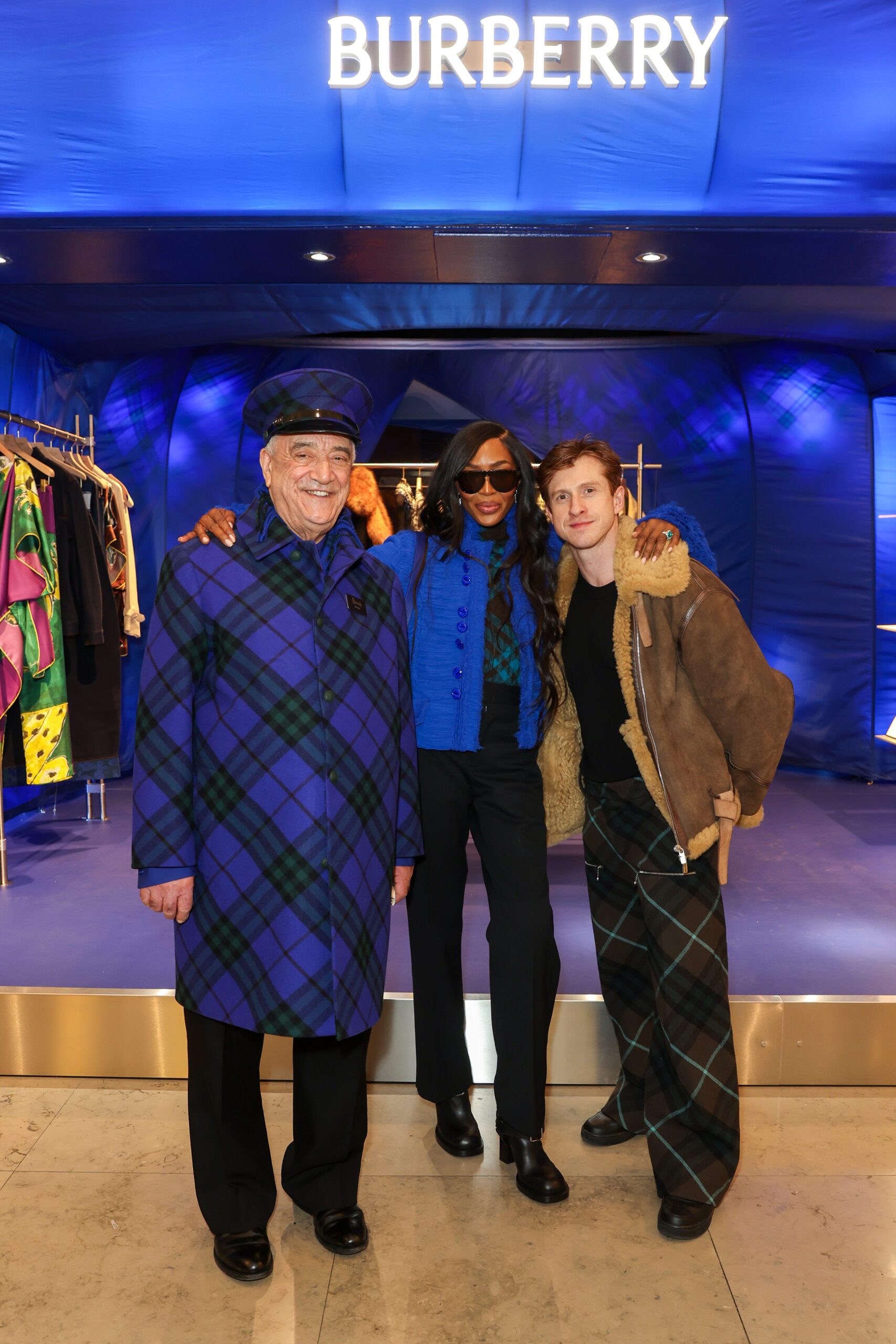 Harrods doorman, Naomi Campbell and Burberry Chief Creative Officer Daniel Lee pose together at the Burberry takeover of Harrods event, with a vibrant Burberry blue backdrop.