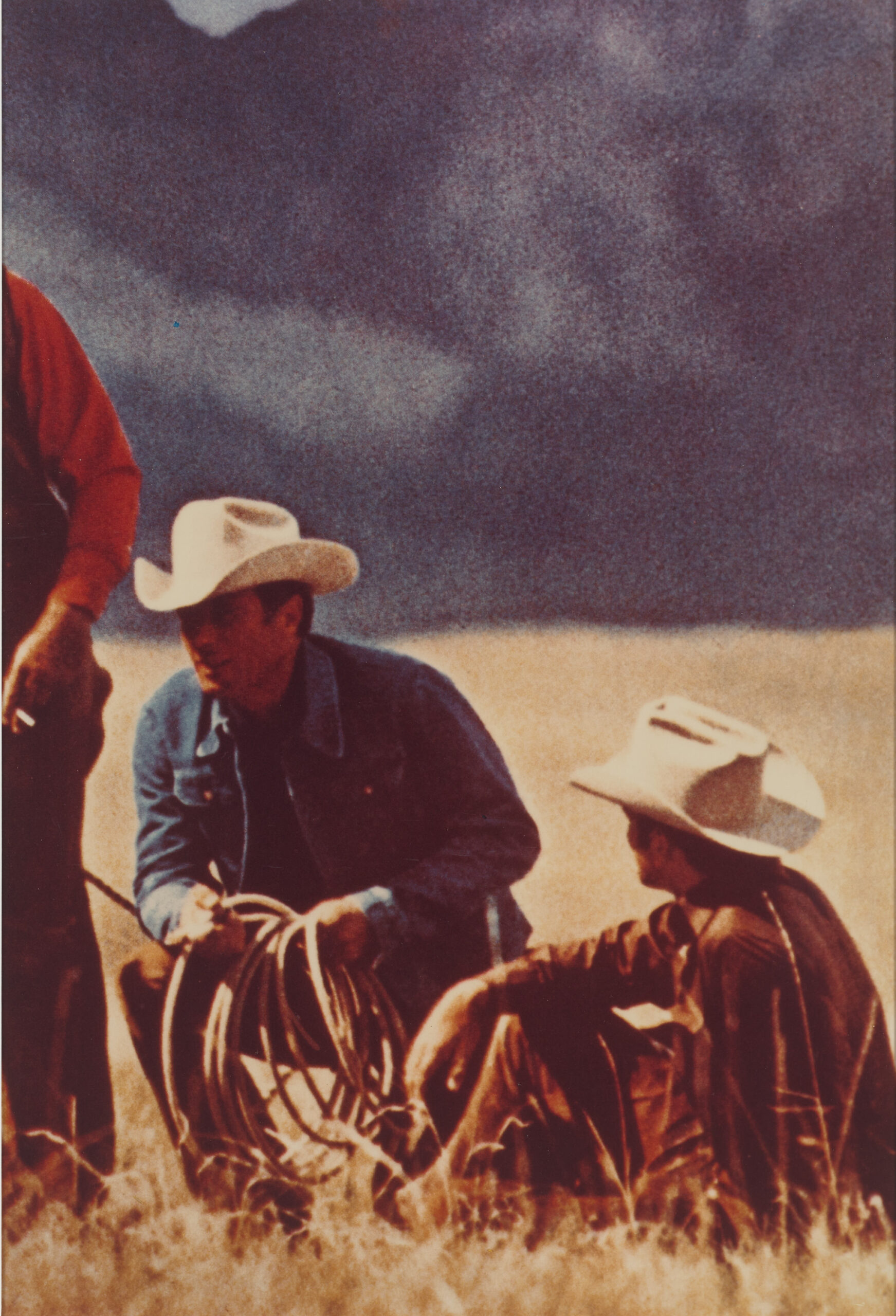 This is an artwork of an Ektacolor photograph depicting three cowboys in a field under a stormy sky. The central figure is seated and holding a lasso, flanked by another cowboy kneeling and a standing figure partially cropped from the frame.
