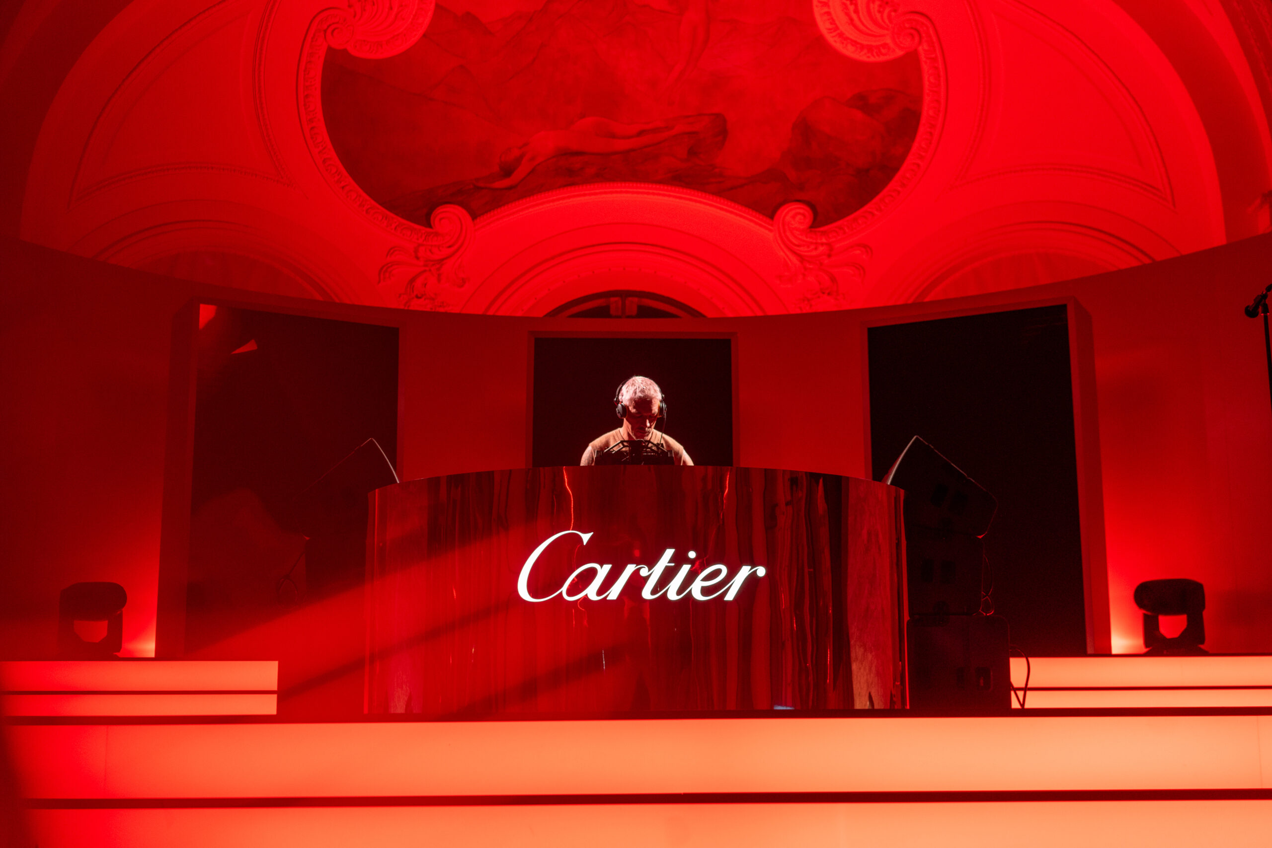 A DJ performs at a Cartier event, surrounded by red lighting and the brand's logo prominently displayed on the front of the DJ booth.