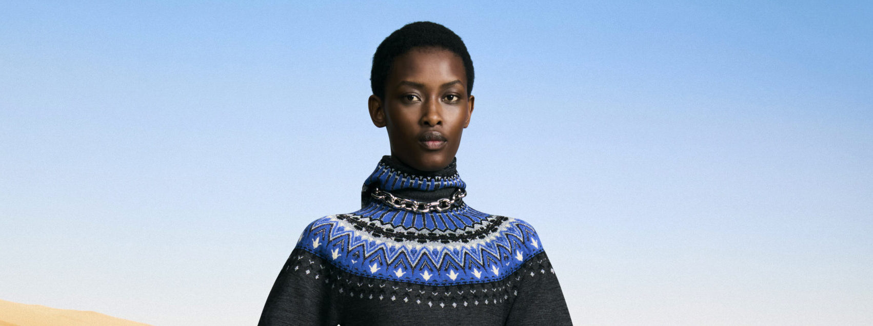 A model poses confidently wearing a Paco Rabanne Pre-fall 2024 ensemble featuring a high-neck sweater adorned with intricate geometric patterns in shades of blue, black, and white, and accented with a stylish metal chain necklace.