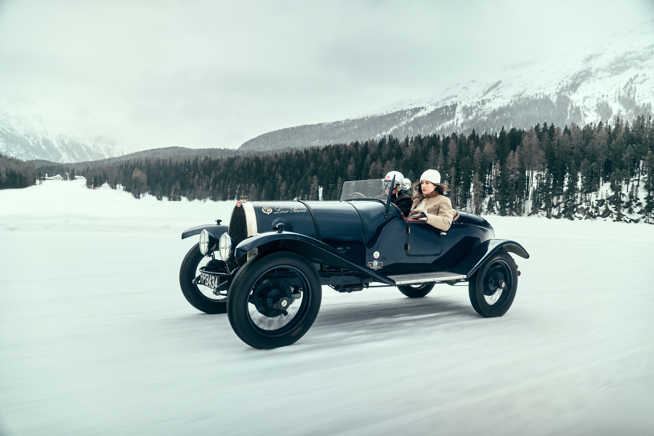 A vintage 1923 Bugatti Brescia T23 16 Valvole in motion on the frozen lake of St. Moritz with a female passenger wearing a white beanie and a beige jacket, both reflecting the luxurious style of Loro Piana.