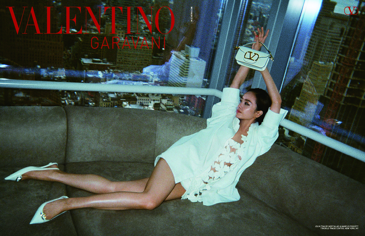 Jolin Tsai reclines on a modern sofa against a window showcasing New York City's skyline. She is elegantly dressed in a white ensemble with delicate cut-out details, emblematic of Valentino's L’École Summer 2024 collection. Her arms are gracefully raised, holding a white VLogo Moon Bag from the Valentino Garavani accessory line, which adds a touch of sophisticated branding to the composition.