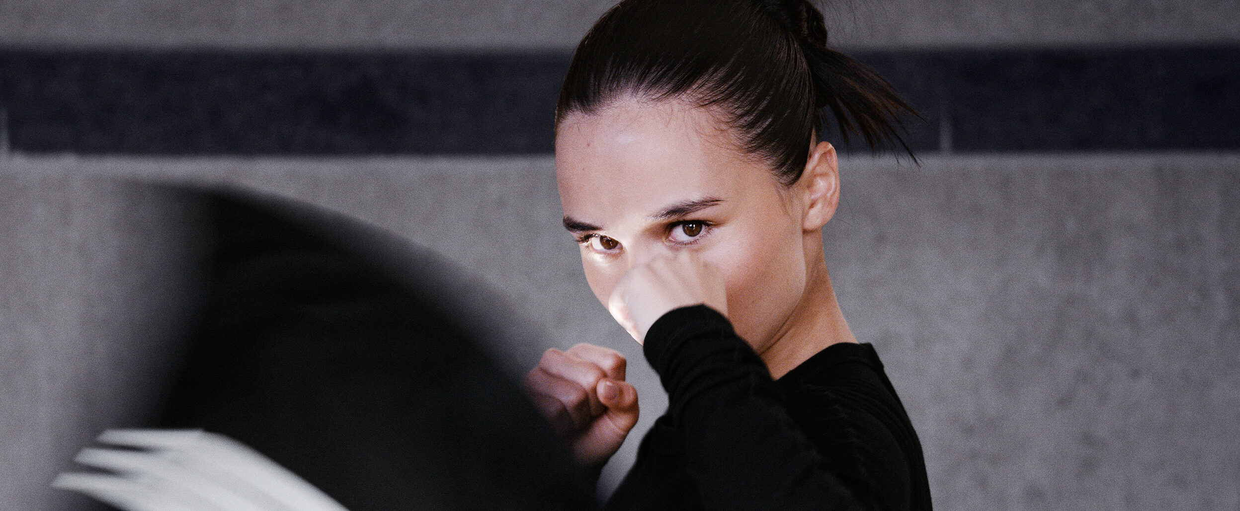 An intense female boxer trains in a black Y-3 sports top with white stripes, her focused gaze directed towards the camera as she throws a punch.