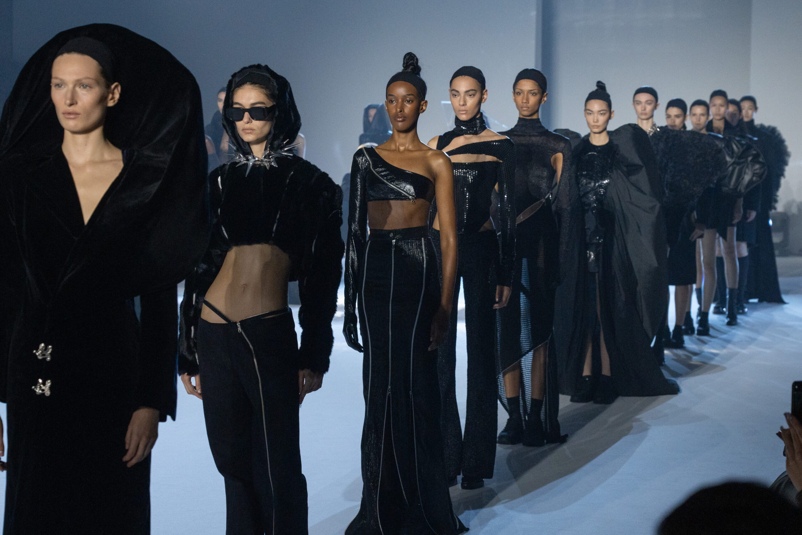 A line of models wearing MAISON YOSHIKI PARIS designs strides down the runway at Milan Fashion Week Fall/Winter 2024/2025. The collection showcases black, tailored pieces with elements of glamor and androgyny. A standout garment includes a dramatic, oversized hooded piece at the forefront.
