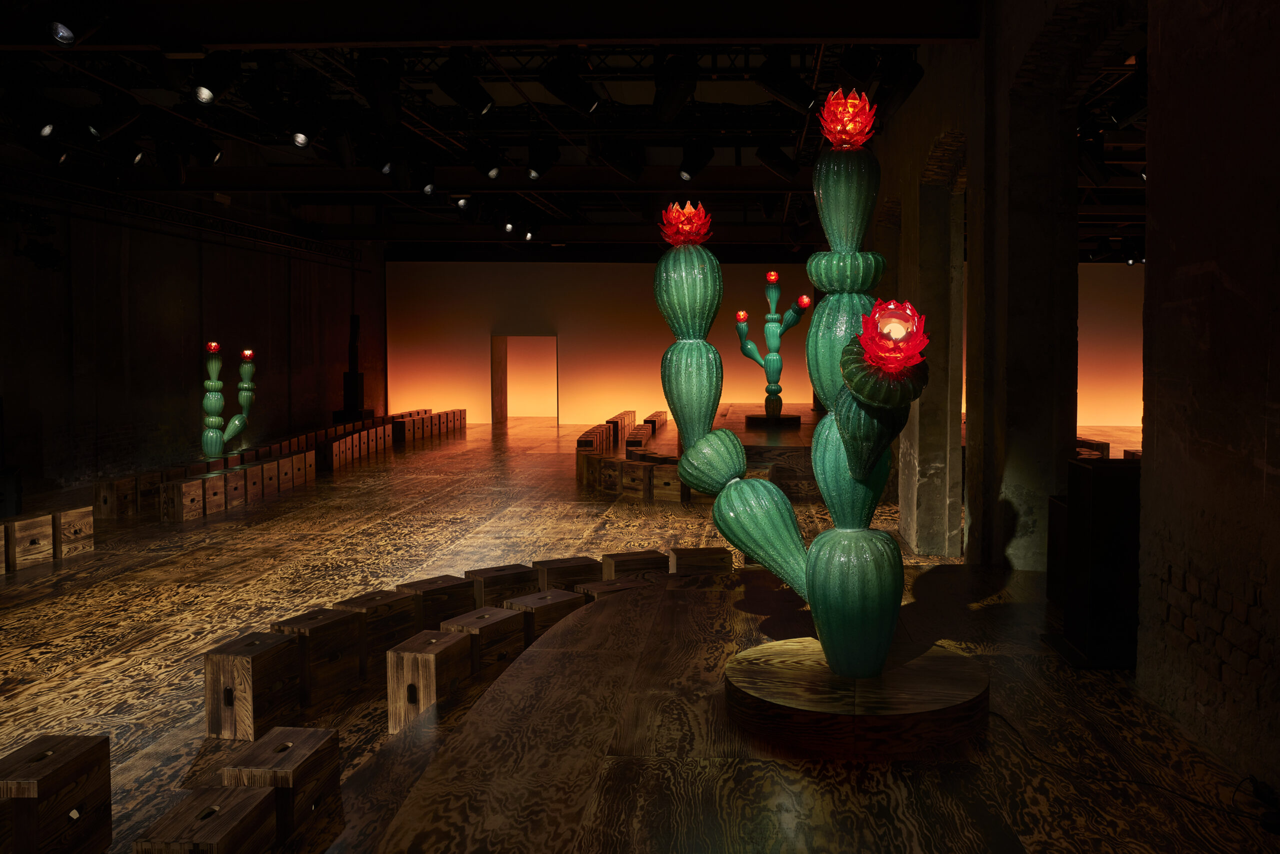 An atmospheric view of Bottega Veneta's Fall/Winter 2024 fashion show venue, featuring artistic cactus sculptures with bright red blooms. The room is dimly lit, highlighting the sculptures and the warm, fiery backdrop at the end of the runway.