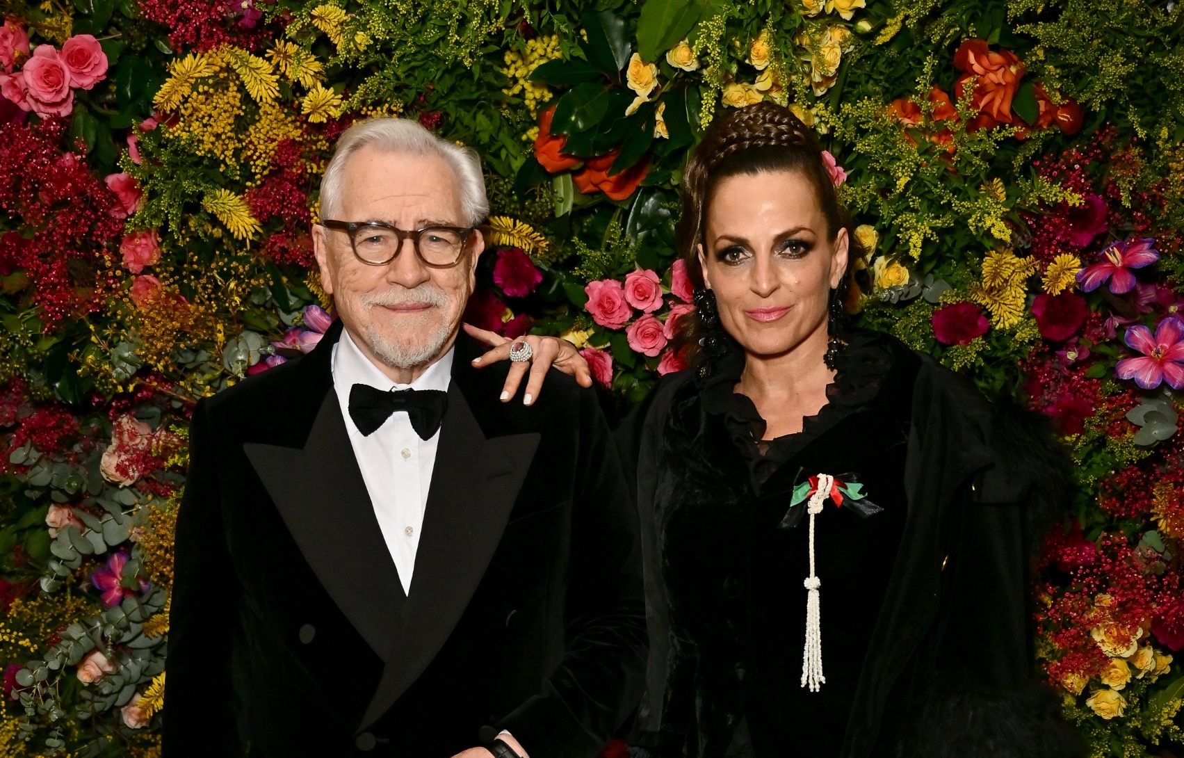 Brian Cox and a woman stand together at the British Vogue Fashion and Film Party 2024. Cox wears a black velvet double-breasted Dunhill jacket, a white shirt, and black velvet Alfred Dunhill Insignia slippers. The woman beside him is in an elegant black ensemble with a distinctive white tassel accessory. They are framed by a vibrant backdrop of assorted flowers.