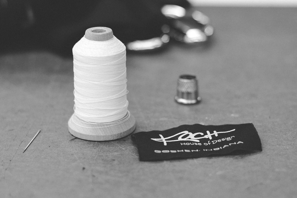 A grayscale image of a sewing setup from Koch House of Design, featuring a spool of white thread, a sewing needle, a thimble, and a black fabric label with "Koch House of Design" written in white cursive and block letters.