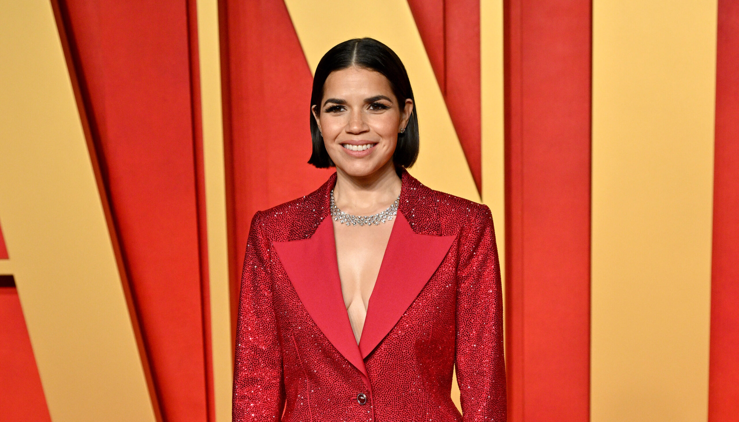 America Ferrera stands with confidence on the Vanity Fair Oscar Party red carpet, wearing a vibrant red, sparkling Versace jacket and black bottoms, accessorized with sheer black stockings and high platform heels.