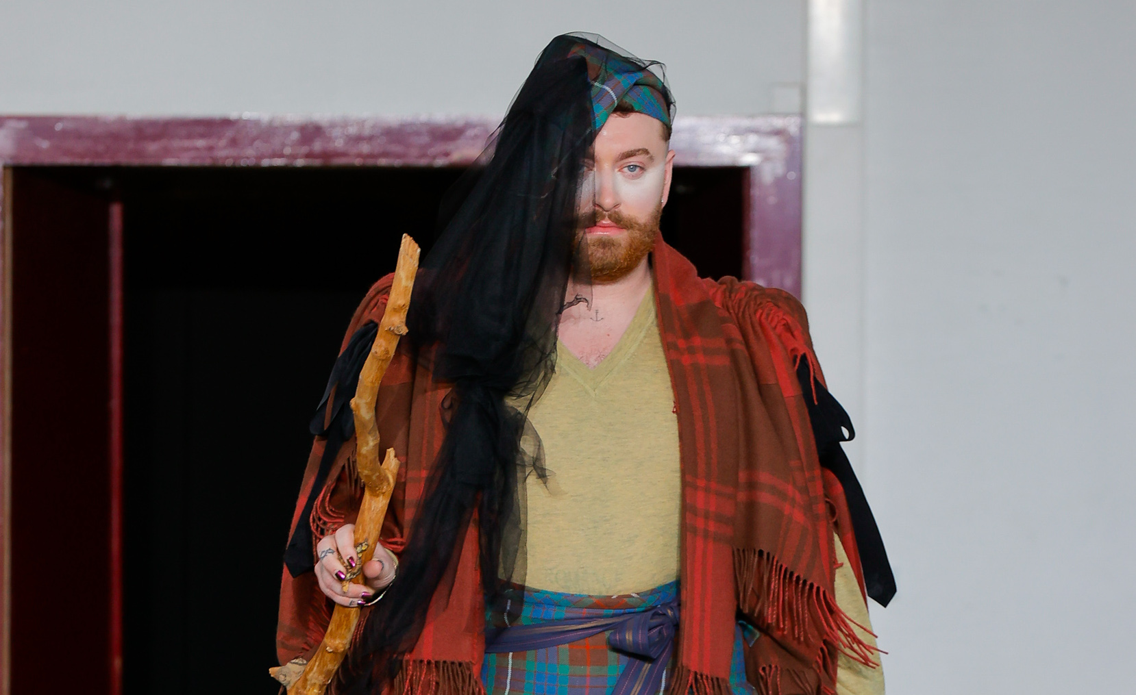 Sam Smith strides confidently on the runway in a vibrant, eclectic ensemble featuring plaid patterns and a rustic staff, at the Andreas Kronthaler for Vivienne Westwood AW24 show.