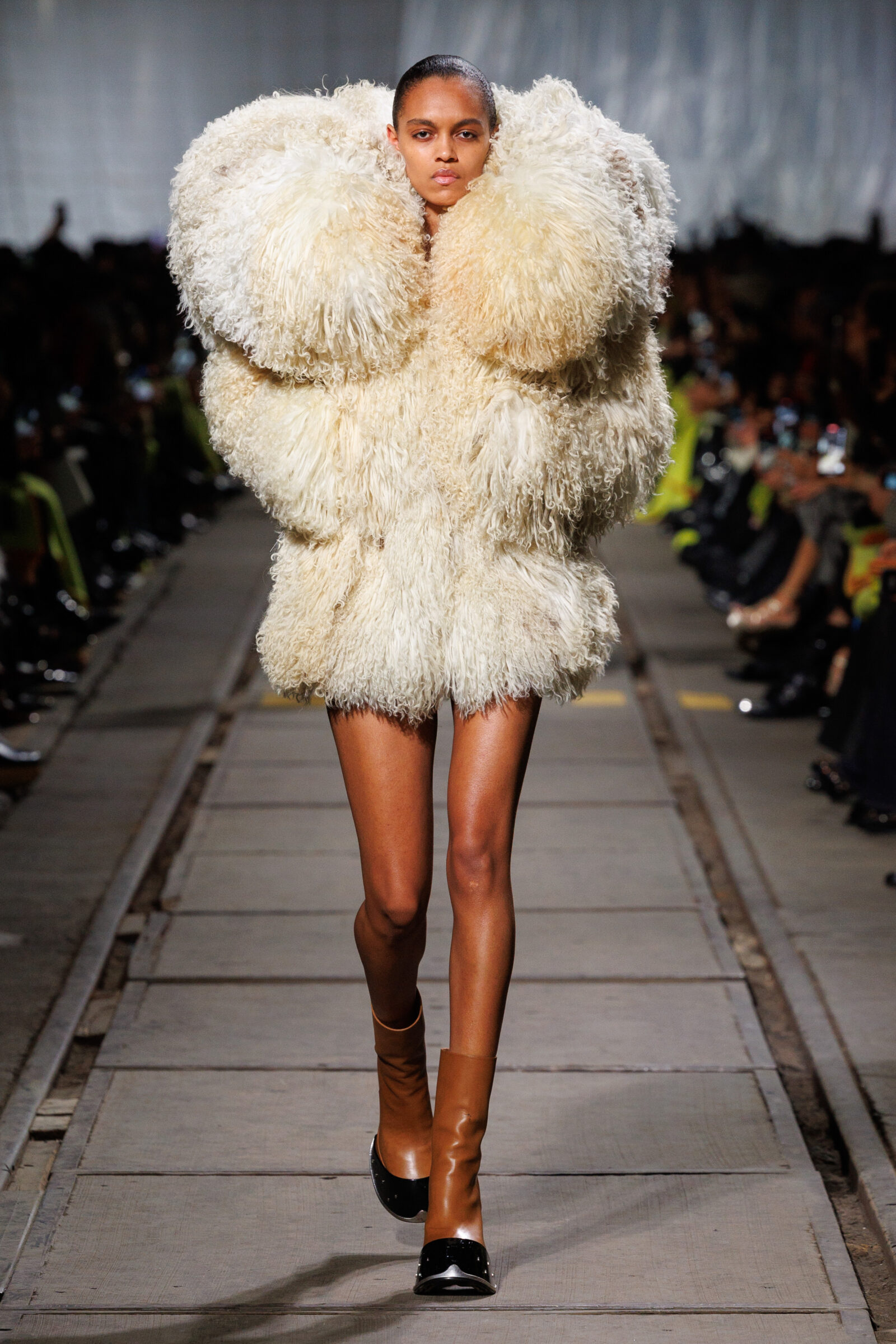 A model commands the runway in an opulent Alexander McQueen shearling coat with exaggerated padded collars, paired with sleek brown leather boots, during the Autumn Winter 2024 show.