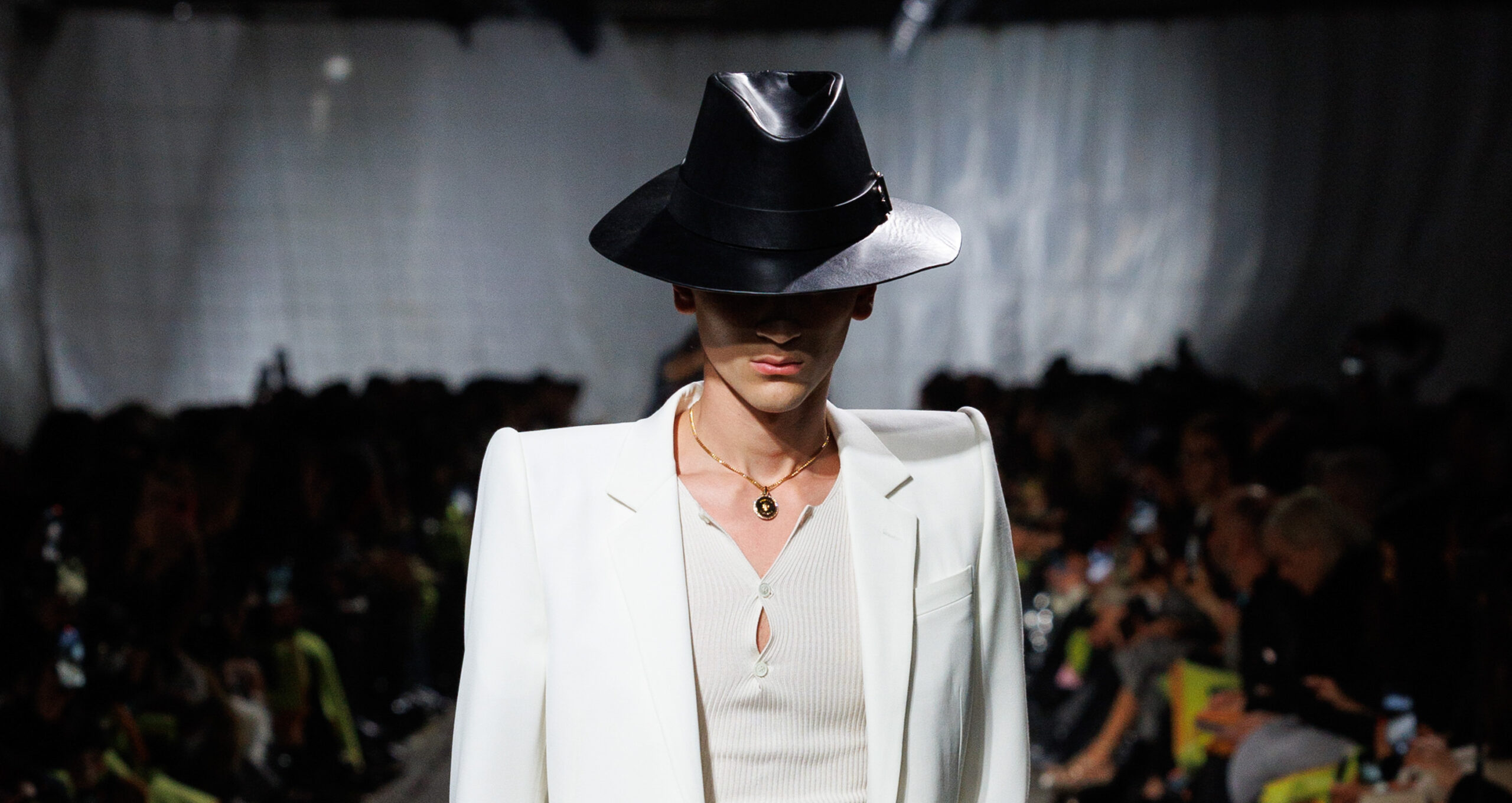 A model walks the runway in an Alexander McQueen Autumn Winter 2024 collection ensemble, featuring a white tailored coat over a fitted knit piece, complemented by a wide-brimmed black hat and a simple gold necklace, against the backdrop of a packed fashion show audience.