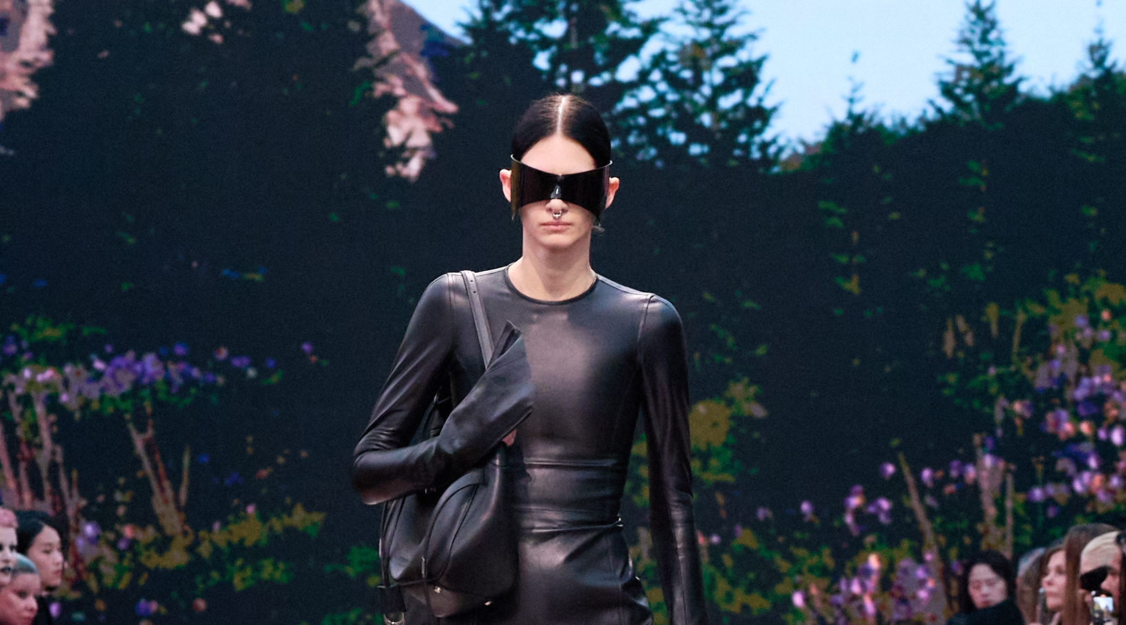 A model is seen walking on the runway clad in an all-black, leather ensemble from the Balenciaga Winter 2024 Ready-to-Wear collection. She is wearing a form-fitting, full-sleeved leather top and carries a coordinating black leather bag. Oversized, sleek sunglasses cover her eyes, adding an edge to the monochromatic outfit.