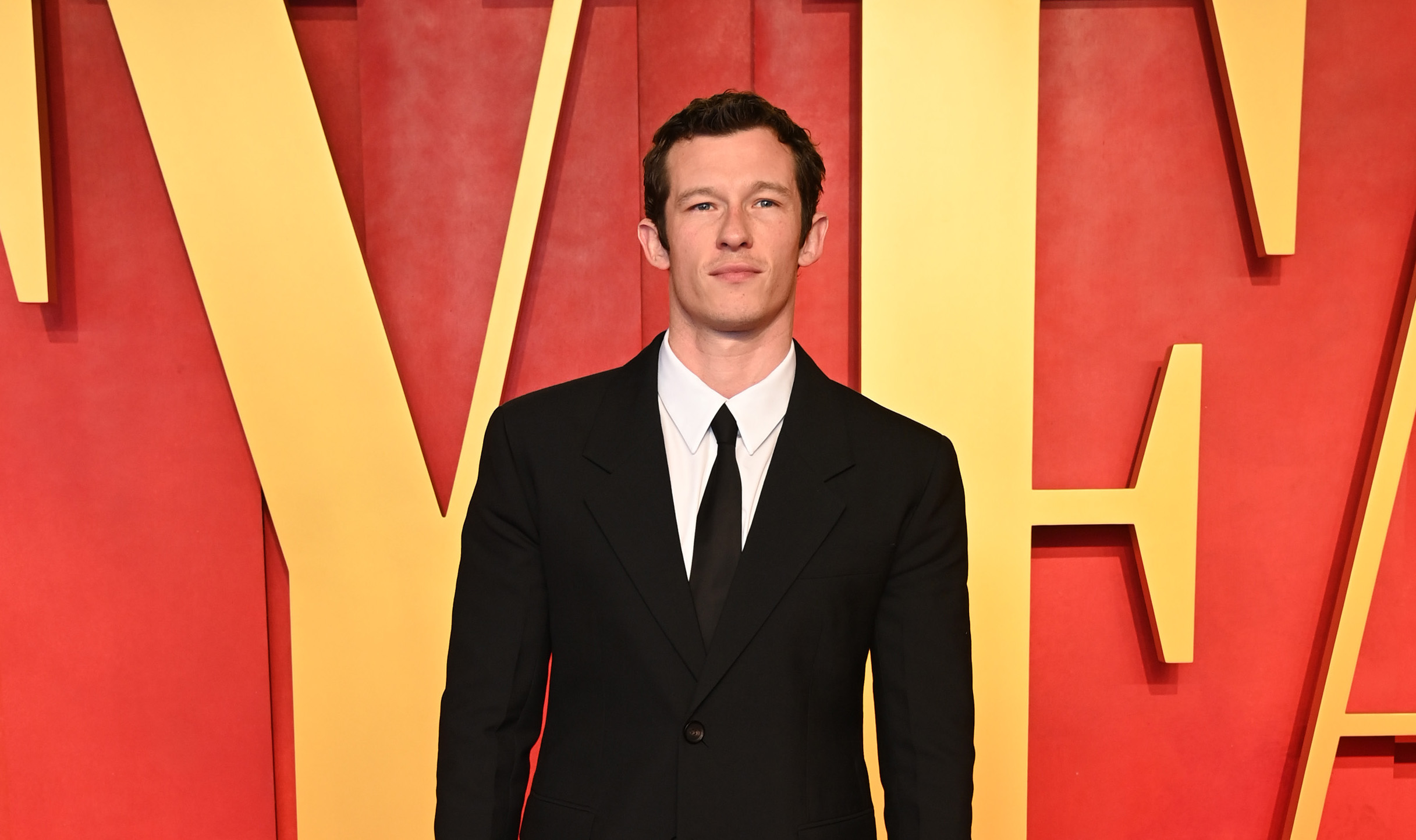 Callum Turner is present at the 2024 Vanity Fair Oscar Party, wearing a Ferragamo single-breasted black Natte wool suit, complemented by a white cotton poplin shirt and black tie, finished with black calfskin ankle boots.