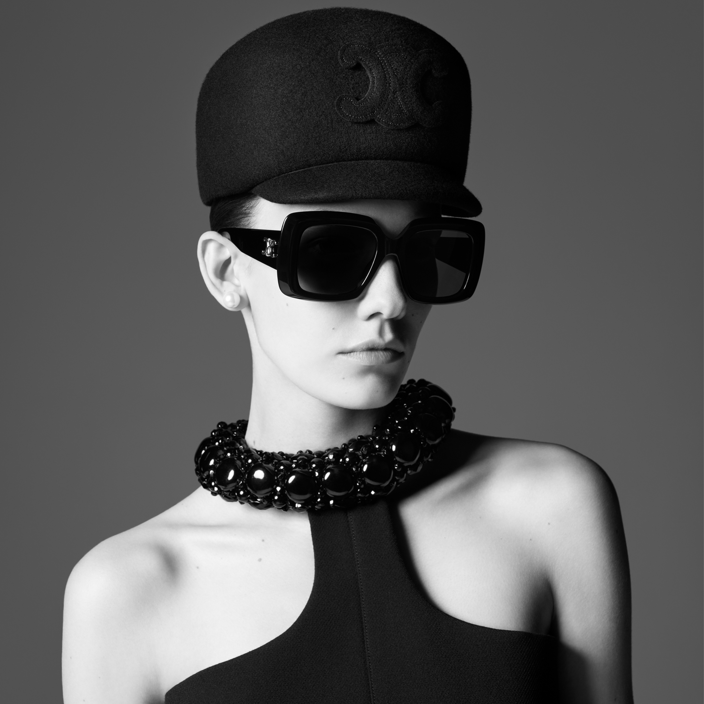 A black and white image of a model with a striking black military cap and oversized sunglasses, featuring a bold beaded necklace against a grey backdrop.