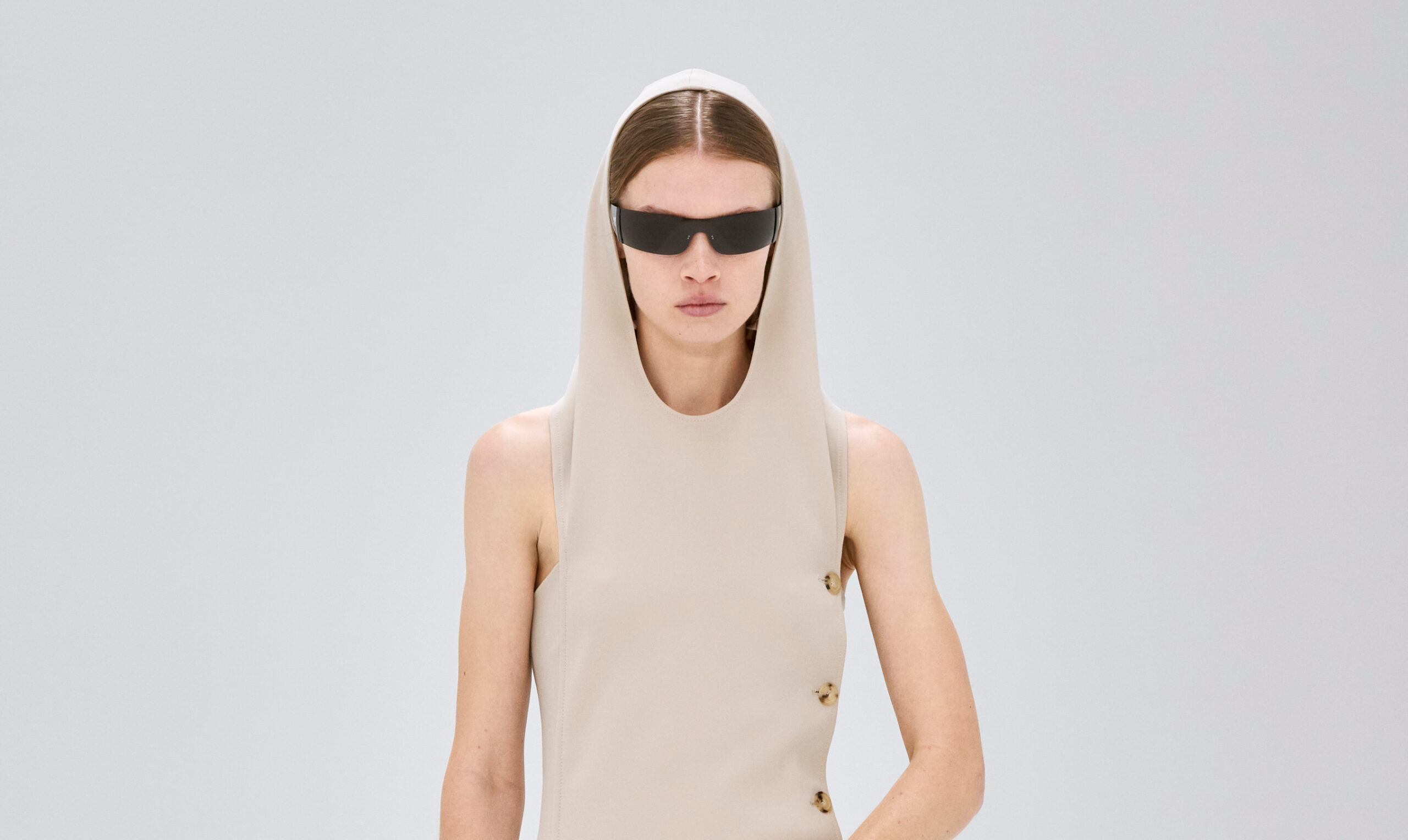 A model from the Courrèges Fall/Winter 2024 collection at Paris Fashion Week wears an asymmetric beige sleeveless top with a draped hood and side button details, paired with black wraparound sunglasses.