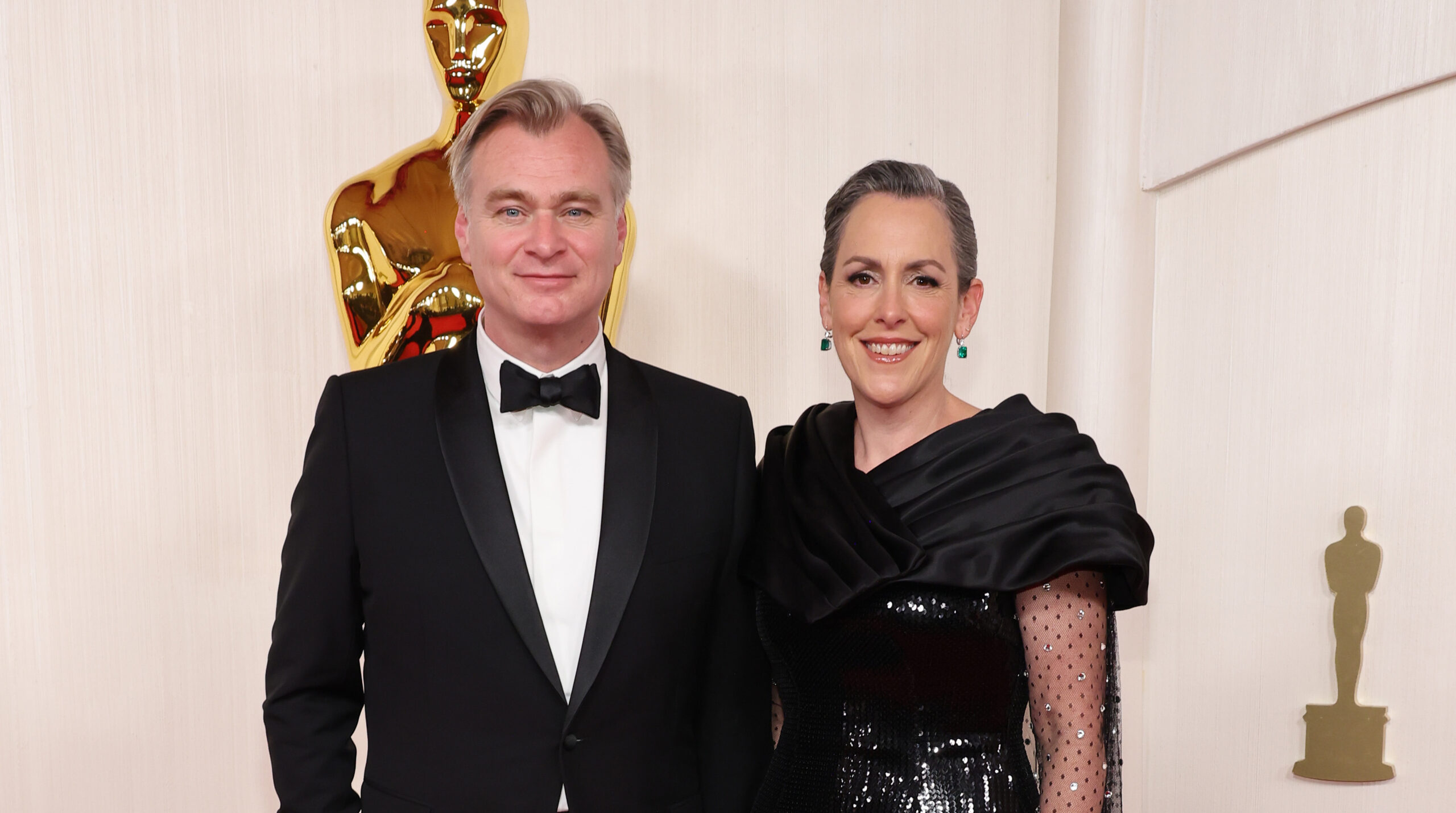 Christopher Nolan and Emma Thomas posing together on the red carpet at the 96th Annual Academy Awards, with Nolan in a classic black Dior tuxedo and Thomas in an elegant black gown with sequin and sheer detailing.