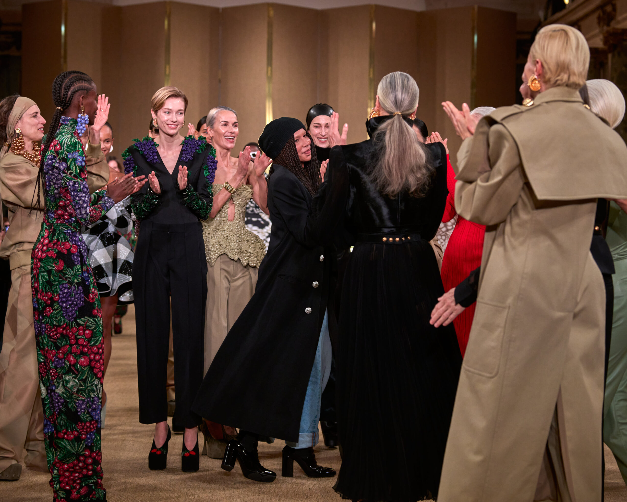 A group of people surround Olivier Rousteing, stand in an indoor setting, clapping and facing a central figure not fully visible in the image. They are stylishly dressed, with the foreground showing various high-fashion garments, like a floral jumpsuit, a black tailored suit, and an embellished green top paired with a black pleated skirt.