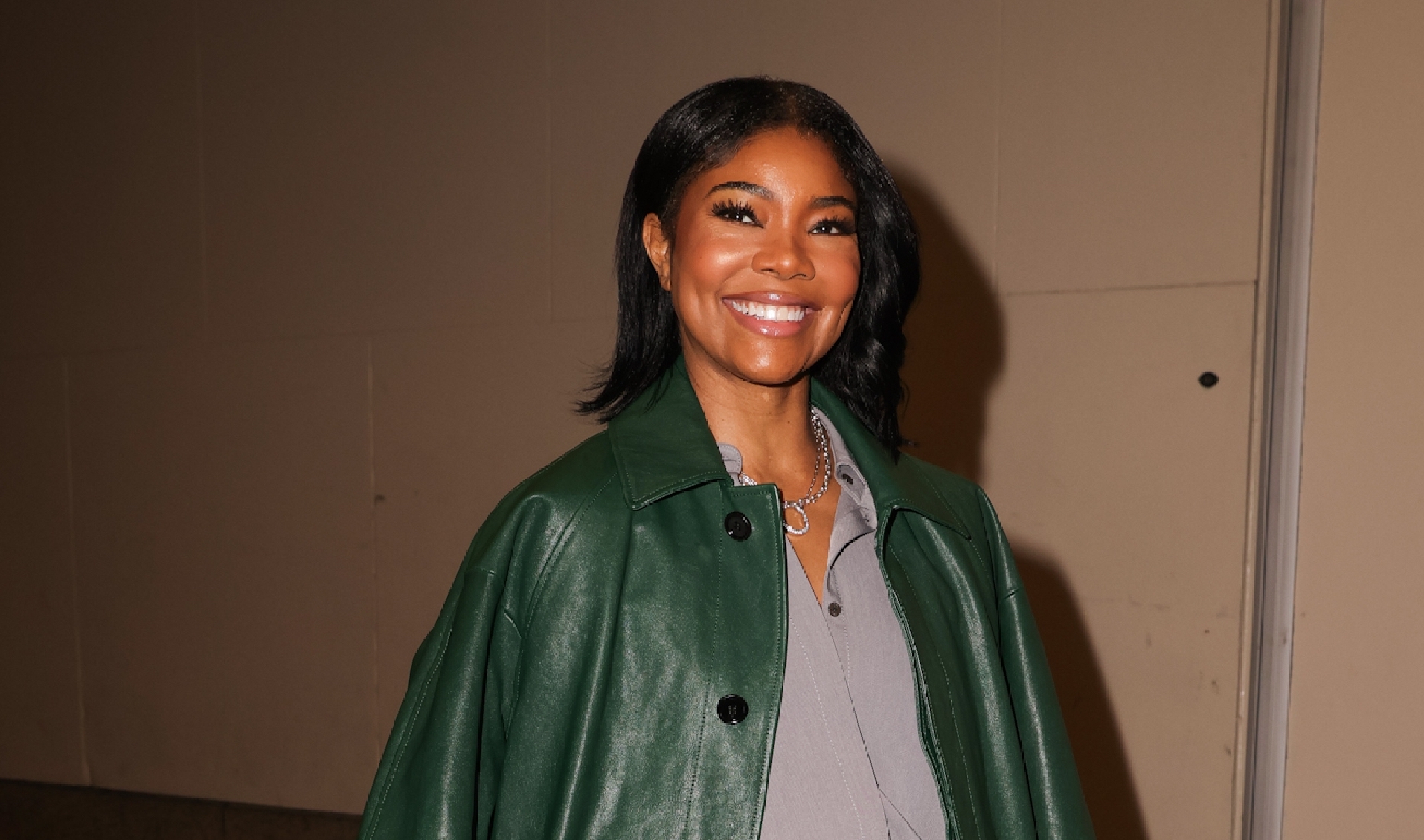 Gabrielle Union is pictured smiling, standing in front of a neutral-toned wall. She is wearing a deep green leather trench coat over a grey top and white mini skirt, accessorized with a black belt and a circular black leather bag. Completing the look are cream-colored leather loafers.