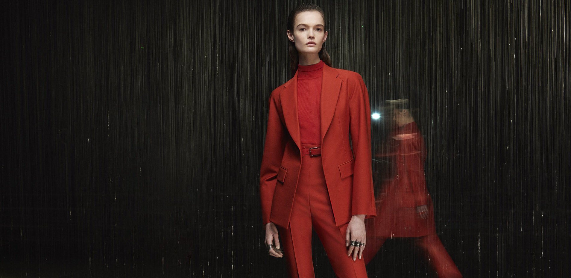 A model dons a tailored red turtleneck ensemble with a structured jacket and belted waist from the Hermès Fall 2024 collection, contrasting with a blurred figure in motion in the background, all set against a rain-like vertical backdrop.