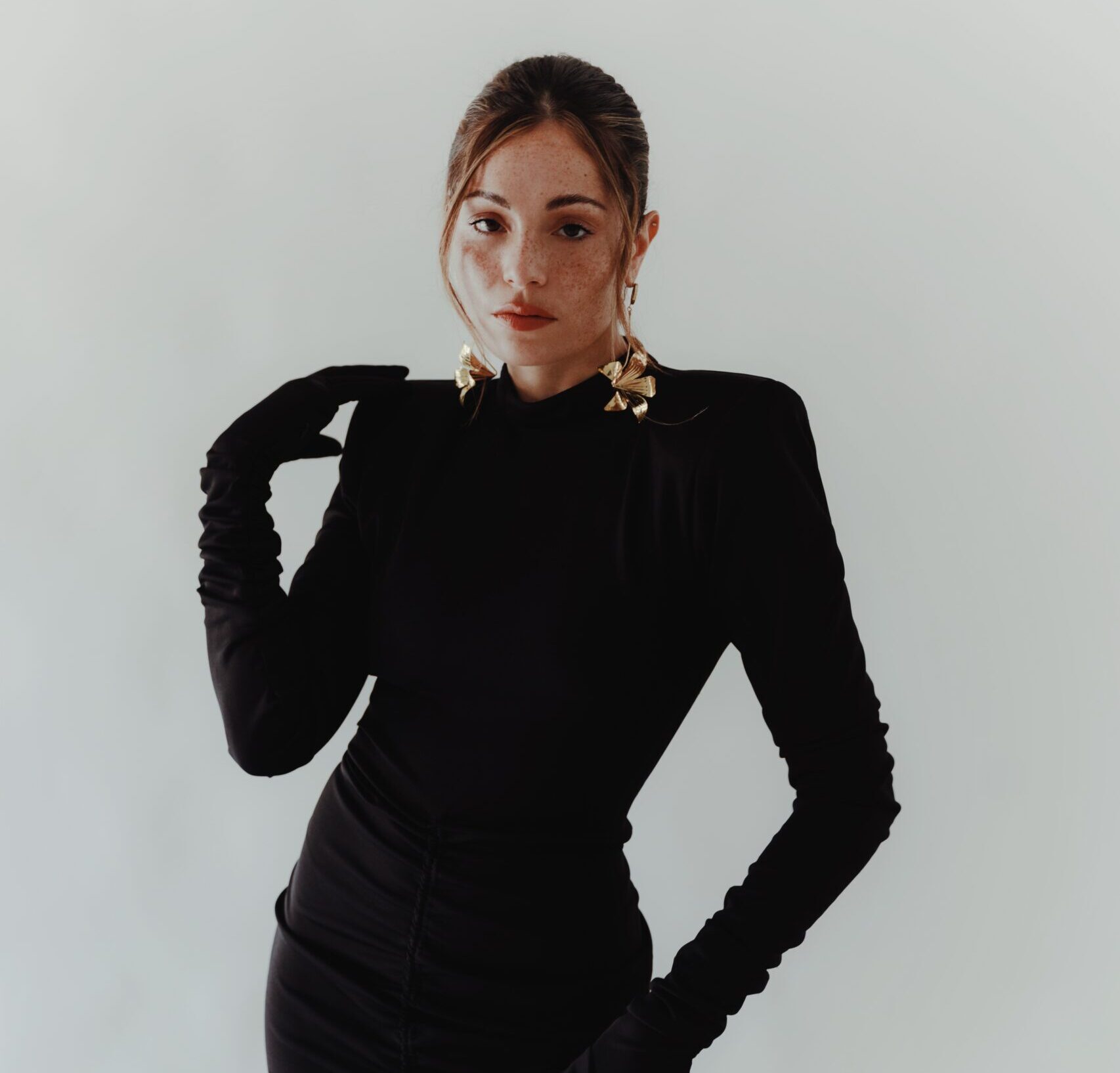 Juliana Aidén Martinez, dressed in an elegant black turtleneck, poses confidently with her hand on her hip. The striking golden earrings add a touch of opulence to her outfit, complementing her self-assured demeanor against a plain white background.