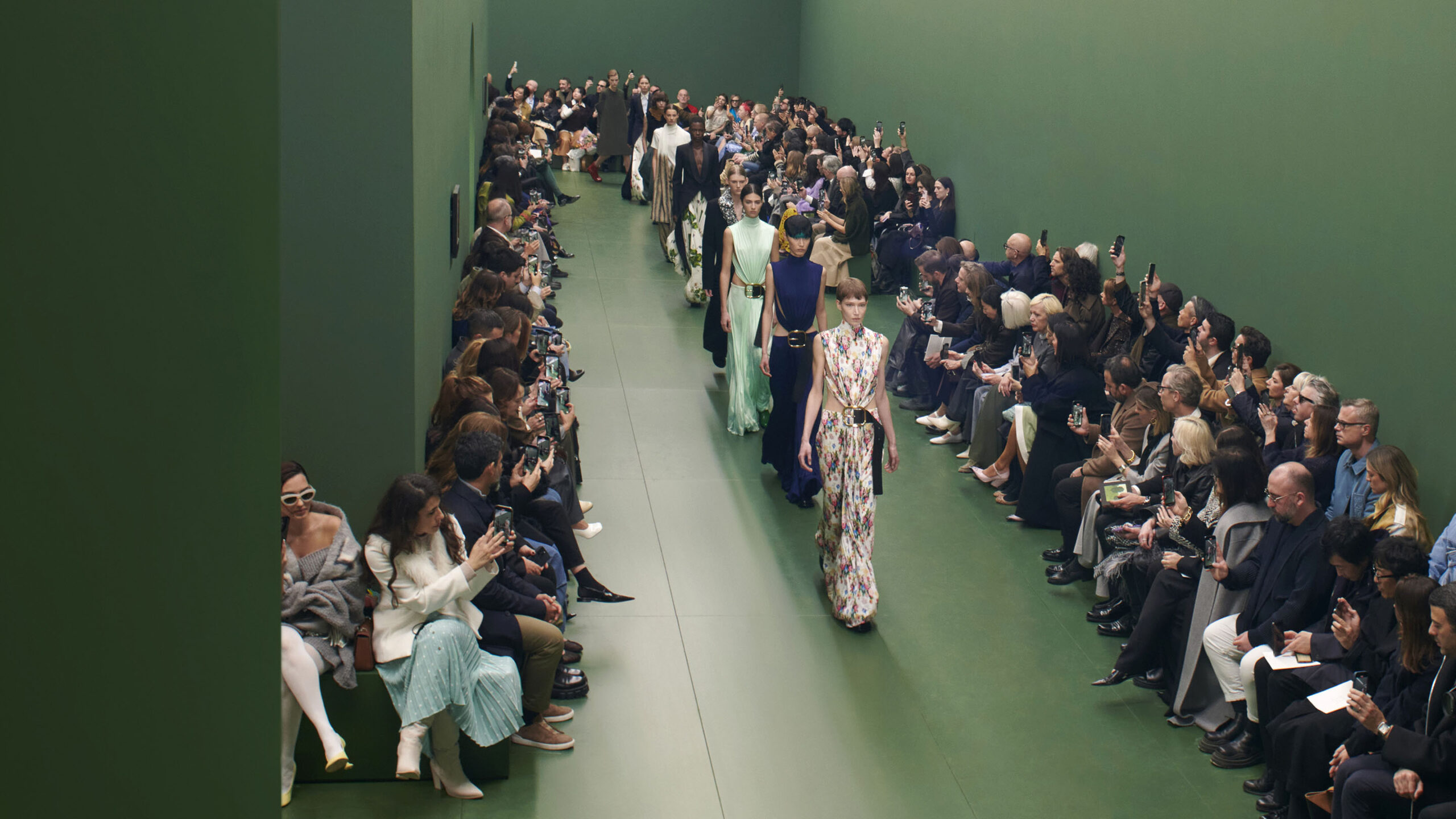 The image displays a runway event during Paris Fashion Week, where models are showcasing the LOEWE Fall/Winter 2024 collection. The setting is characterized by rich green walls, with the audience seated along the sides of the runway, capturing the moment on their phones. Fashion models are adorned in an array of elegant, patterned outfits, walking down the runway which resembles a path through a contemporary Eden, reflecting the artistic influence of Albert York's pastoral landscapes.