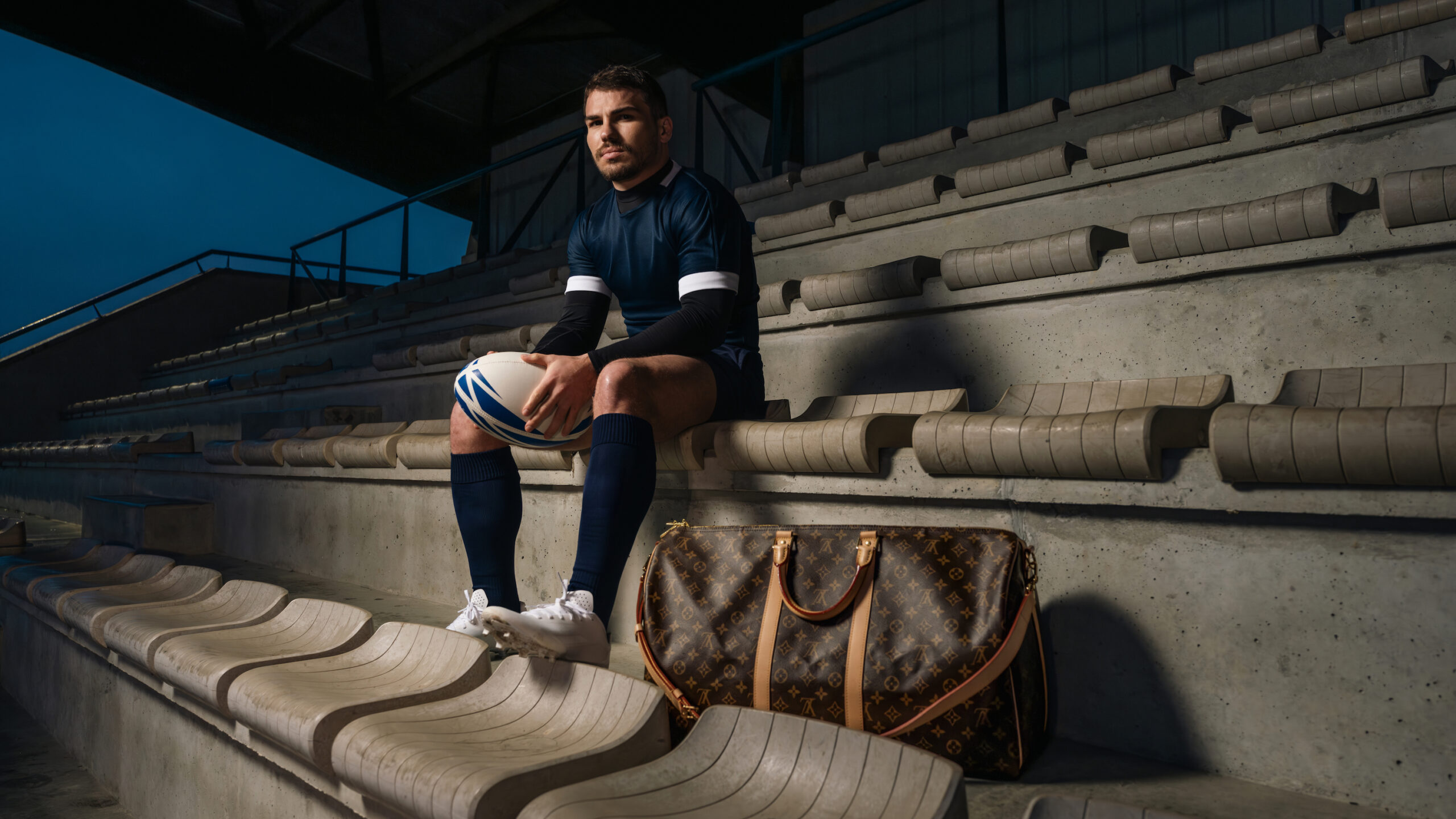 House Ambassador Antoine Dupont, wearing a navy rugby kit, sits on stadium steps with a Louis Vuitton bag beside him and a rugby ball in hand, reflecting the union of sportsmanship and luxury.