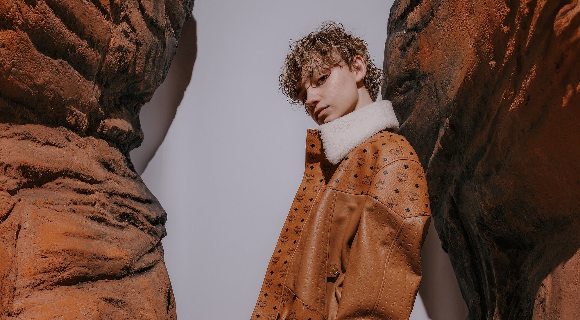 Close-up of a male model wearing an MCM embossed leather jacket with a shearling collar, against a Mars-like rocky backdrop.