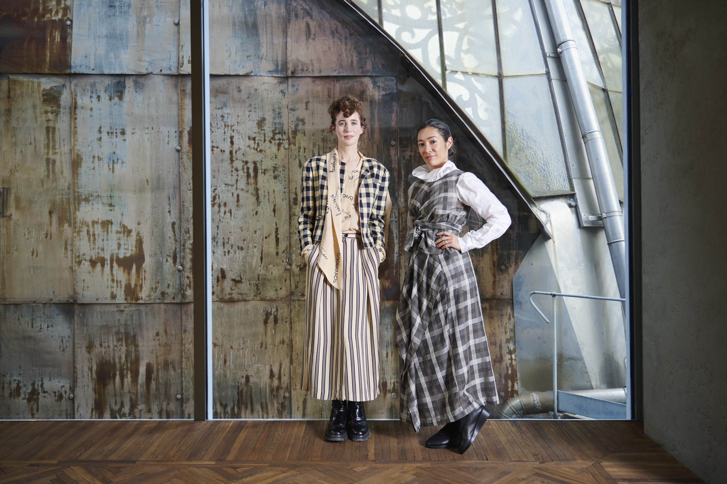 Miranda July and Mia Locks in chic, patterned outfits stand confidently in front of a rustic metal backdrop with an industrial vibe, complementing the strong, artistic atmosphere of the setting.