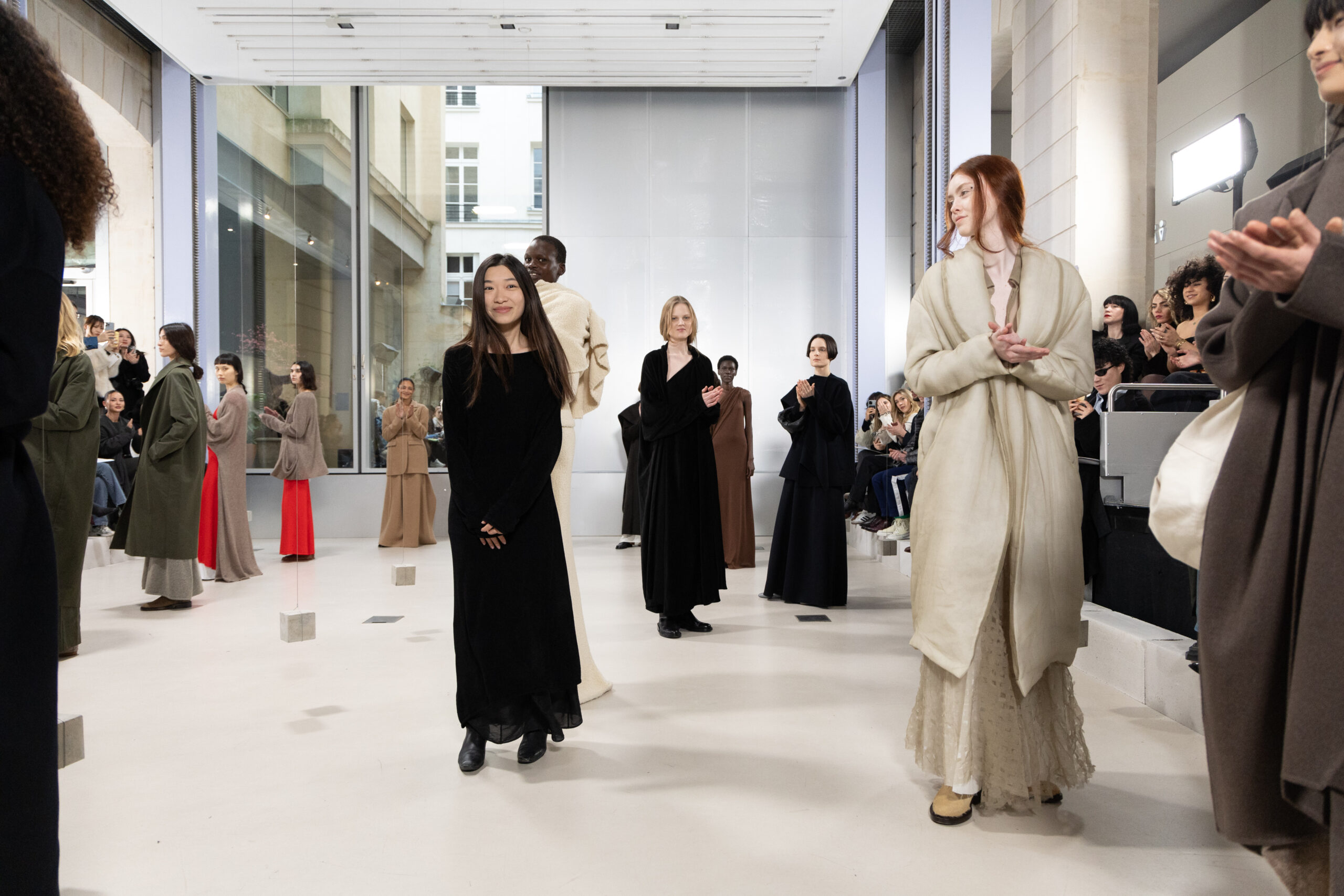 A fashion show finale with models in minimalist, geometric clothing standing in a bright indoor space with large windows, receiving applause from an audience.
