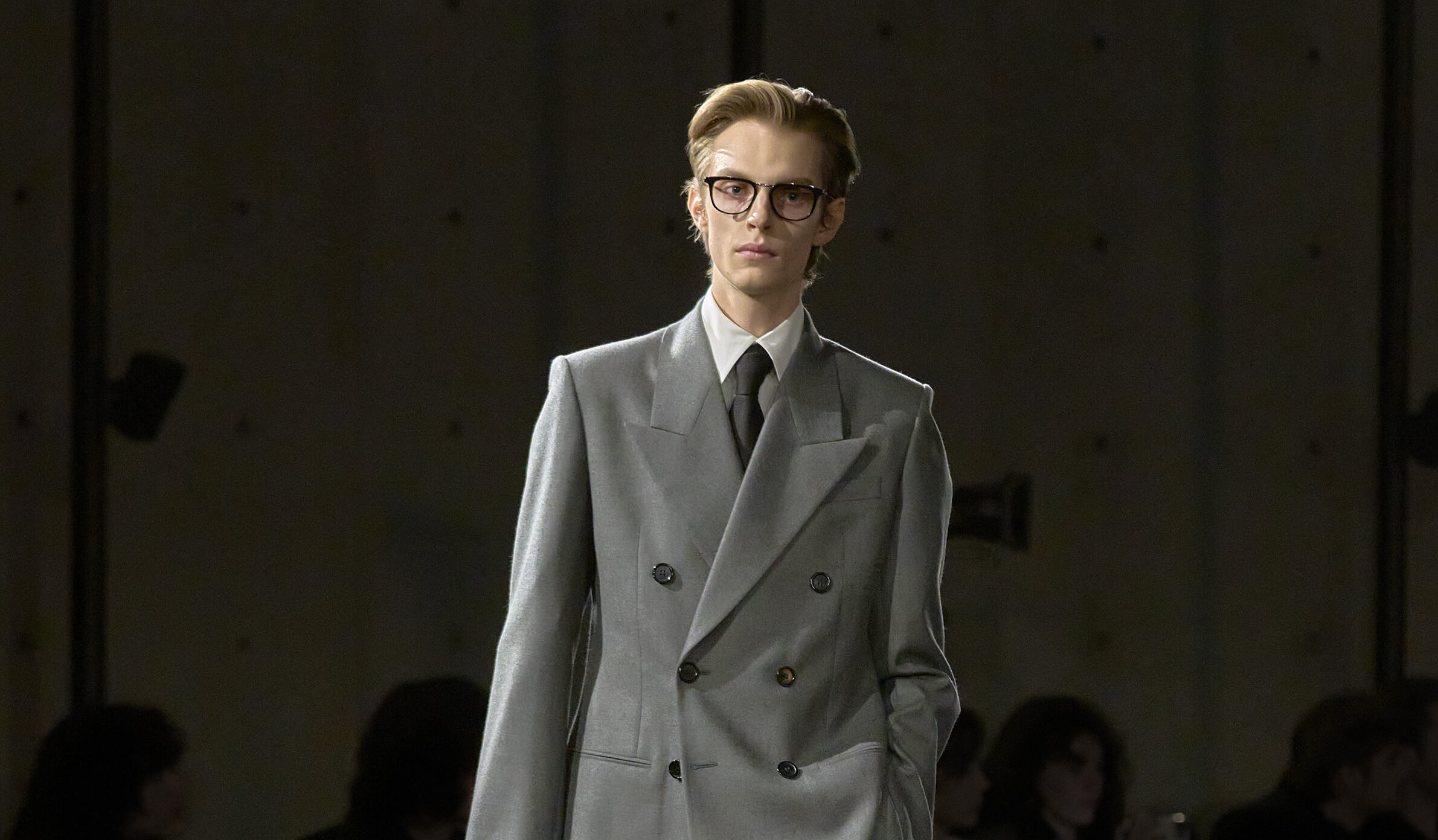 Leon Dame walks down the runway at Saint Laurent's Winter 2024 menswear show, wearing a double-breasted, loose-fitting light grey suit with resolute peak lapels, embodying a modern twist on 1980s power dressing.