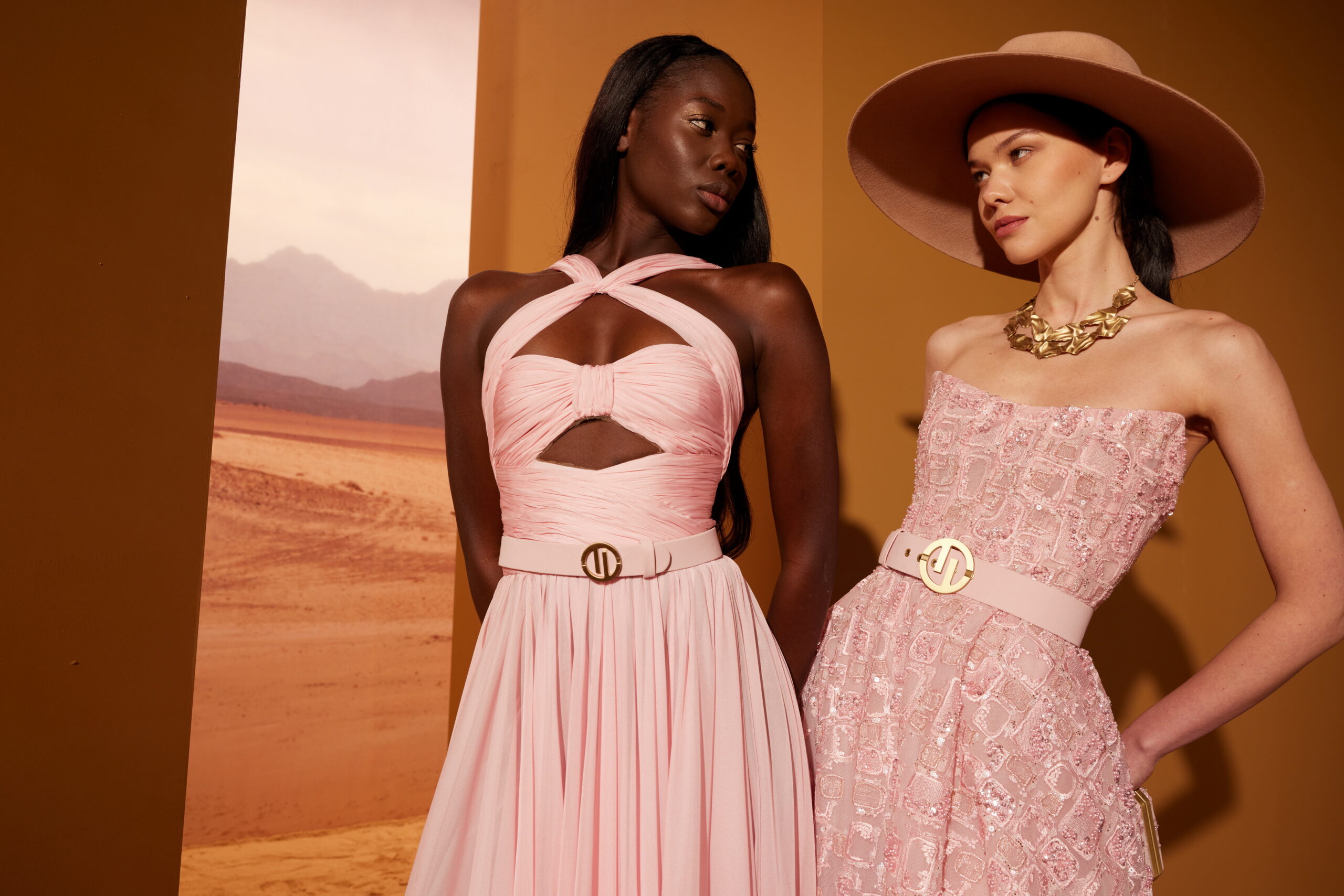 Two models in Tony Ward's "Safari Sunset" collection stand side by side; one model is wearing a pink halter-neck dress with a twist detail and a belted waist, while the other is in a textured, strapless pink dress with a belted cinched waist, complemented by a wide-brimmed hat and a bold gold necklace.